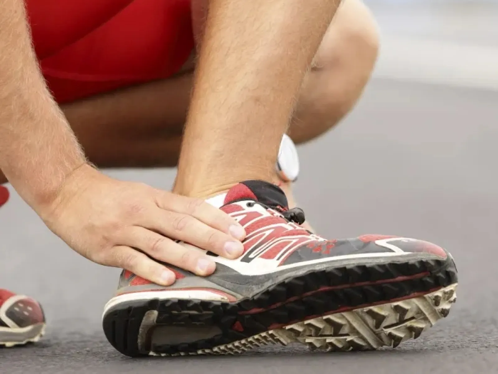 Person wearing red running shoes holding their ankle on an asphalt road, indicating a possible injury or pain.