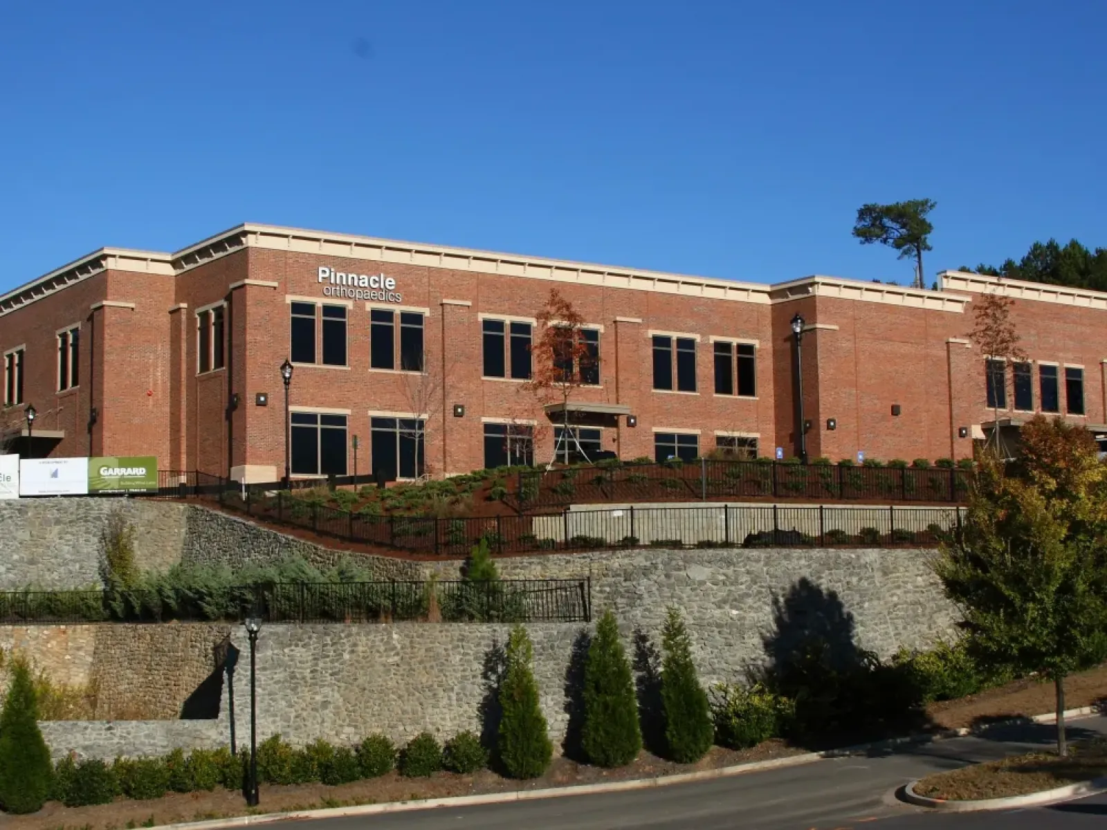 Modern red brick building of Pinnacle Orthopaedics with stone retaining walls and landscaped greenery under a clear blue sky.