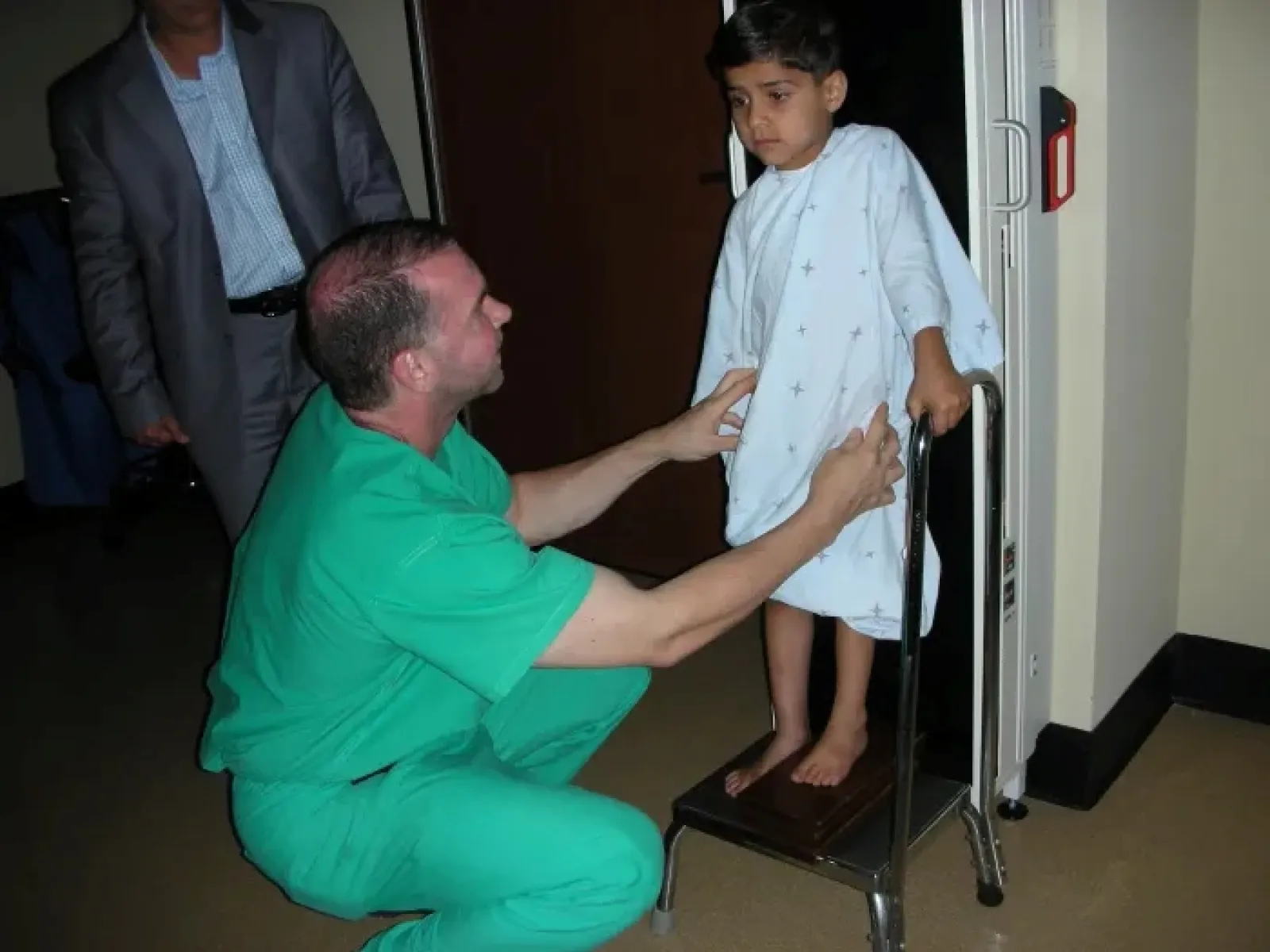 Doctor in green scrubs assisting young boy in hospital gown standing on scale in clinical setting