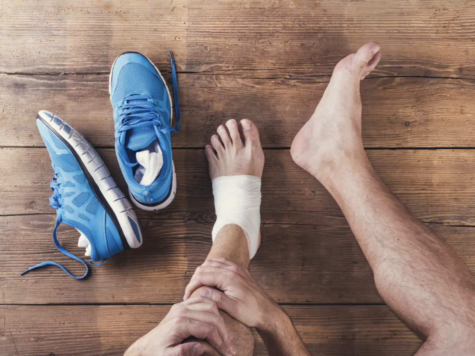 Person with a wrapped ankle sitting on a wooden floor next to blue running shoes and injury advice text.