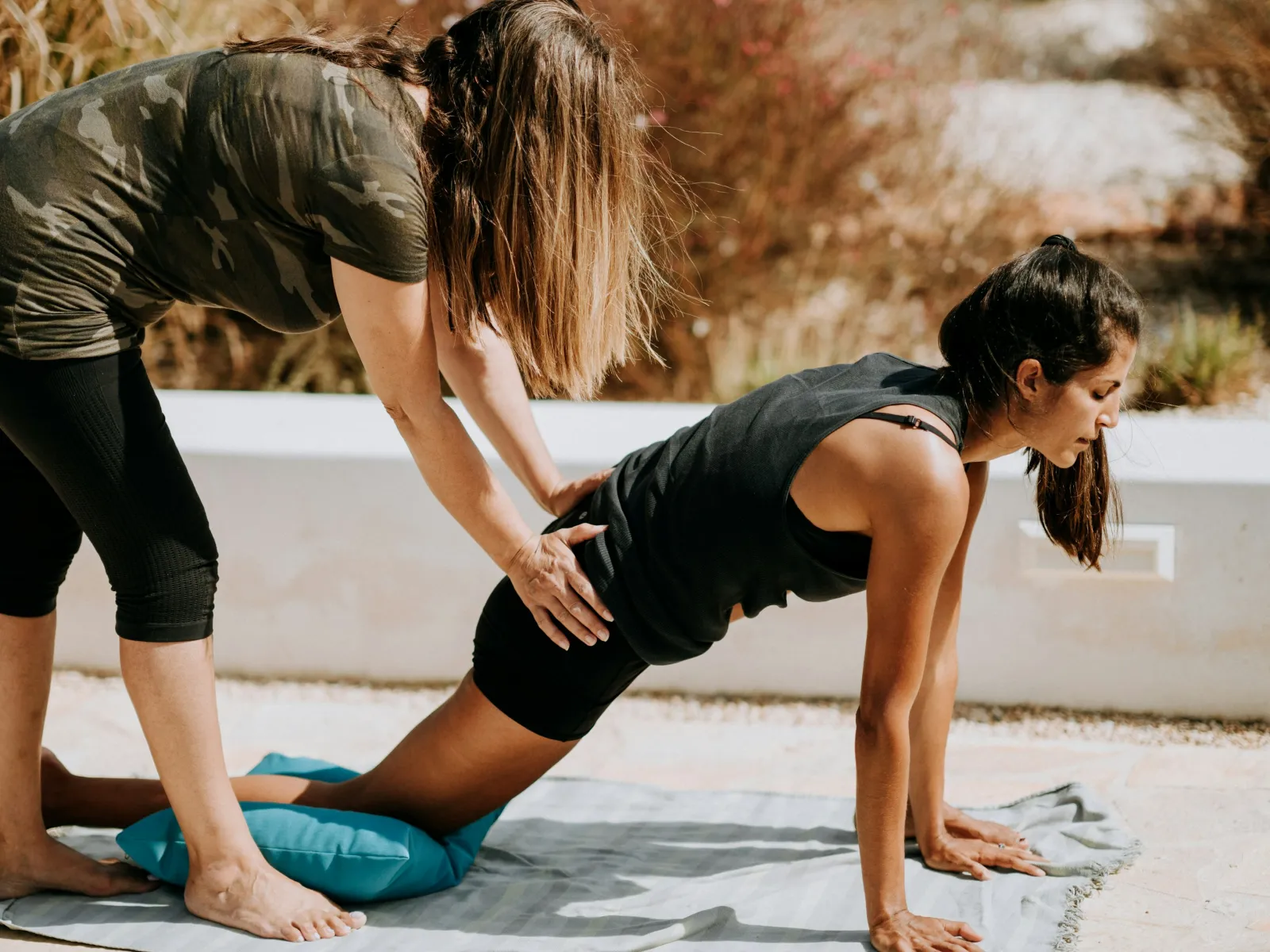 Fitness coach assisting woman in stretching pose outdoors on yoga mat under sun