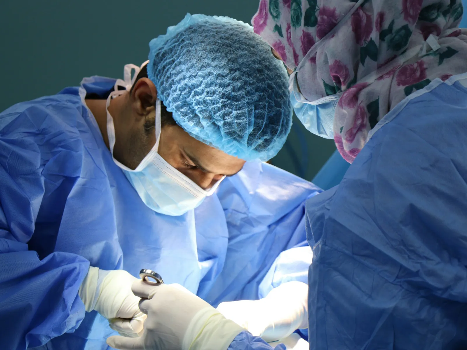 Surgeons in blue surgical gowns and masks performing an operation in a brightly lit operating room.