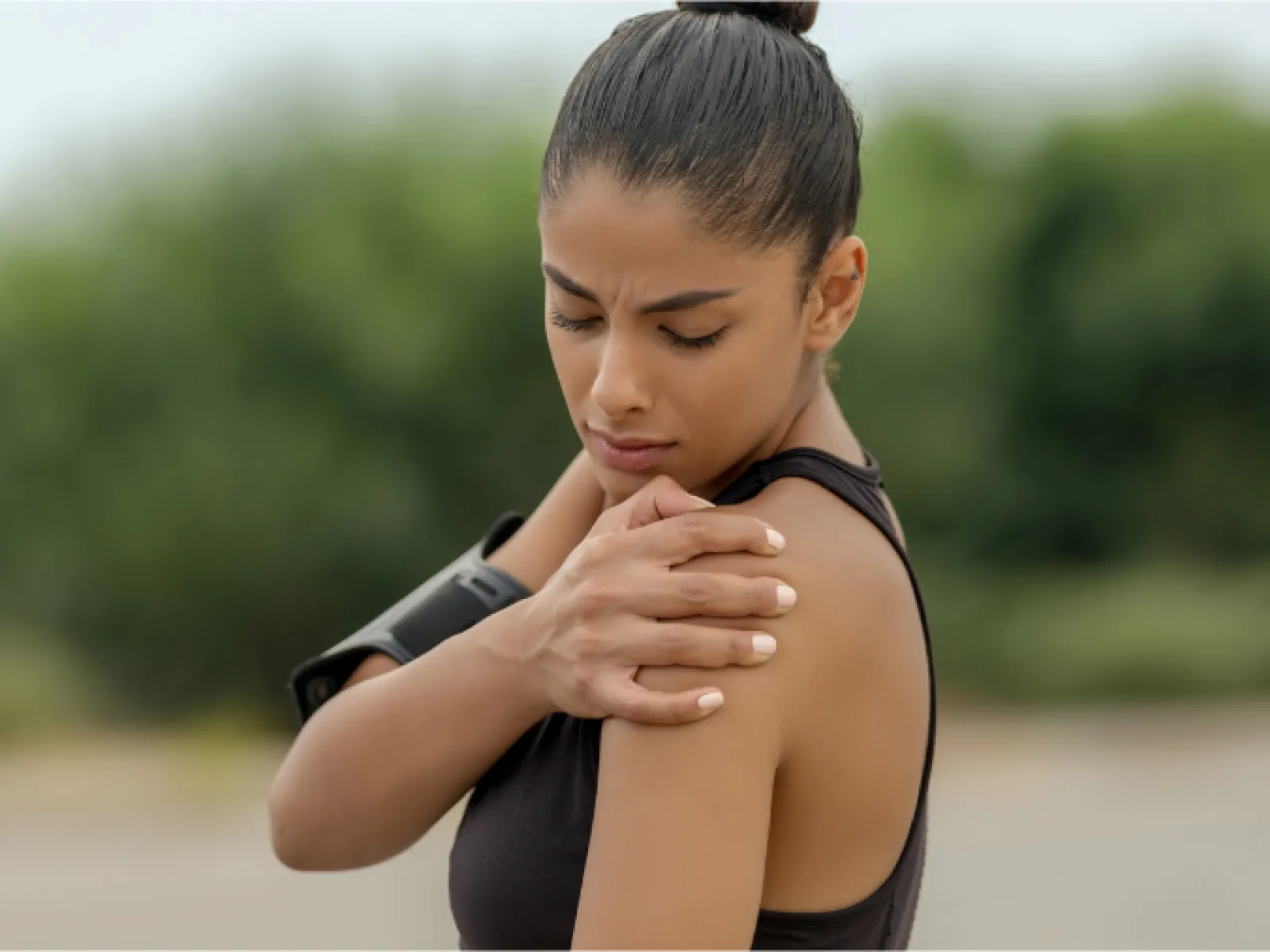 Woman in sportswear holding her painful shoulder outdoors with blurred trees in the background.