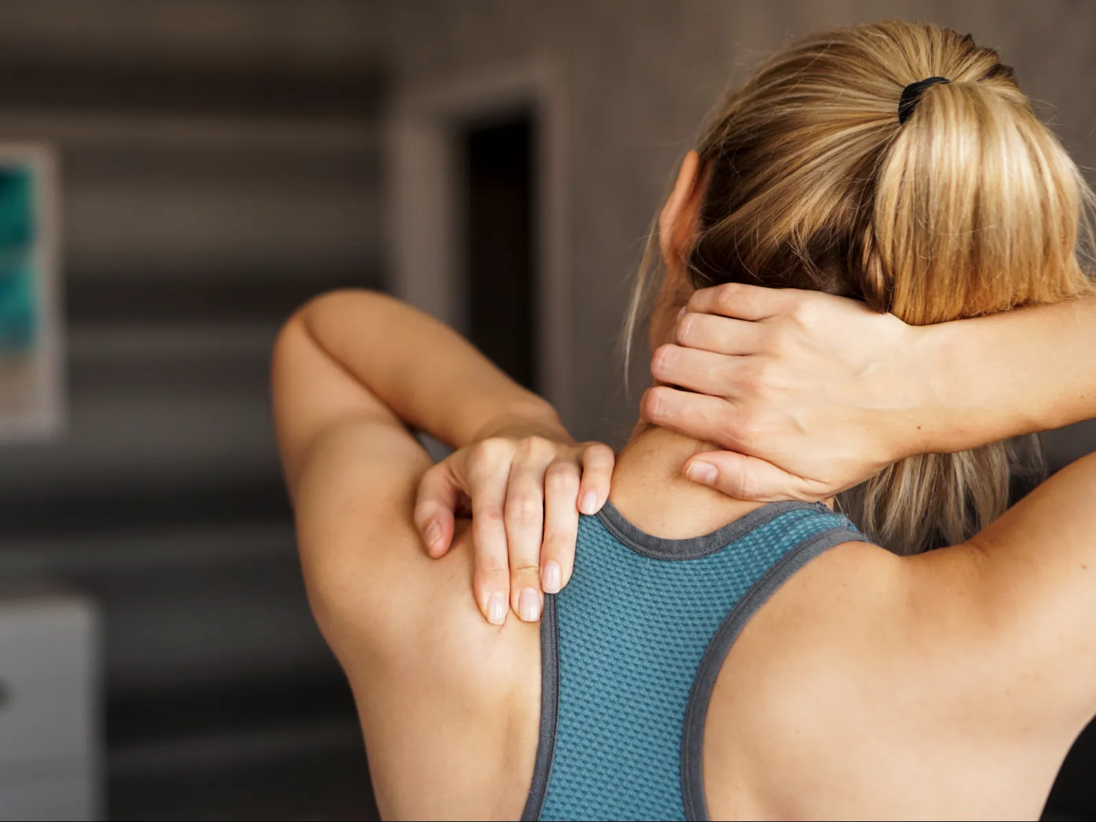 Woman in sportswear massaging her neck and upper back indoors, showing muscle tension relief.