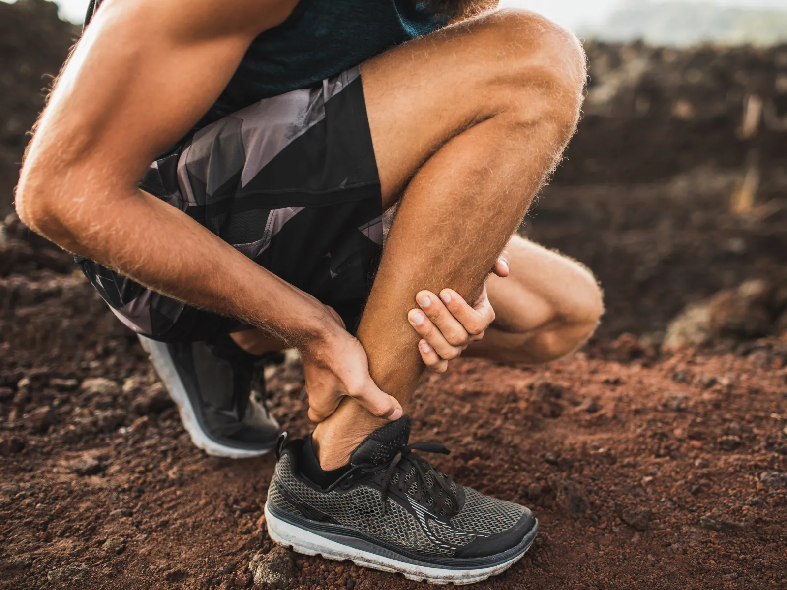 Runner holding ankle in pain on dirt trail wearing black athletic shoes and shorts during outdoor workout.