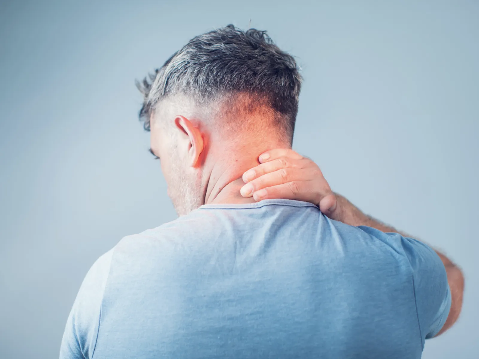 Man in blue shirt holding the back of his neck, showing discomfort or pain against a plain background.
