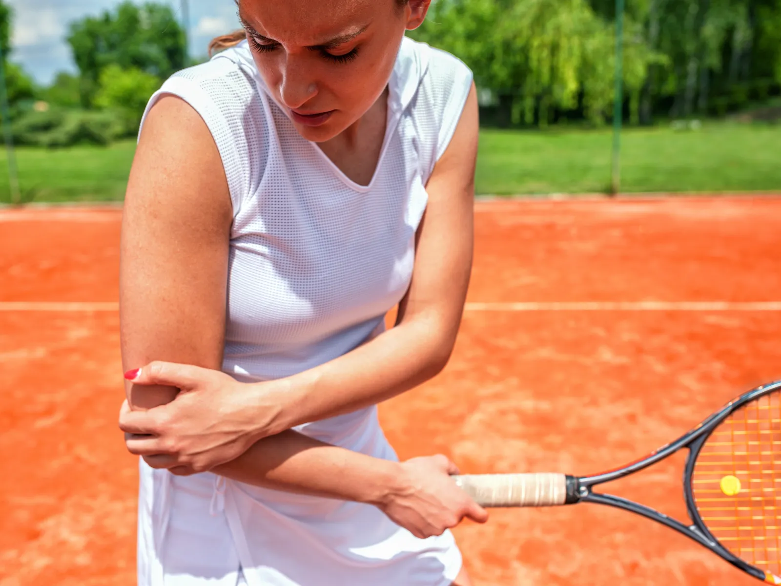 Female tennis player holding her elbow in pain on an outdoor clay court under a sunny sky