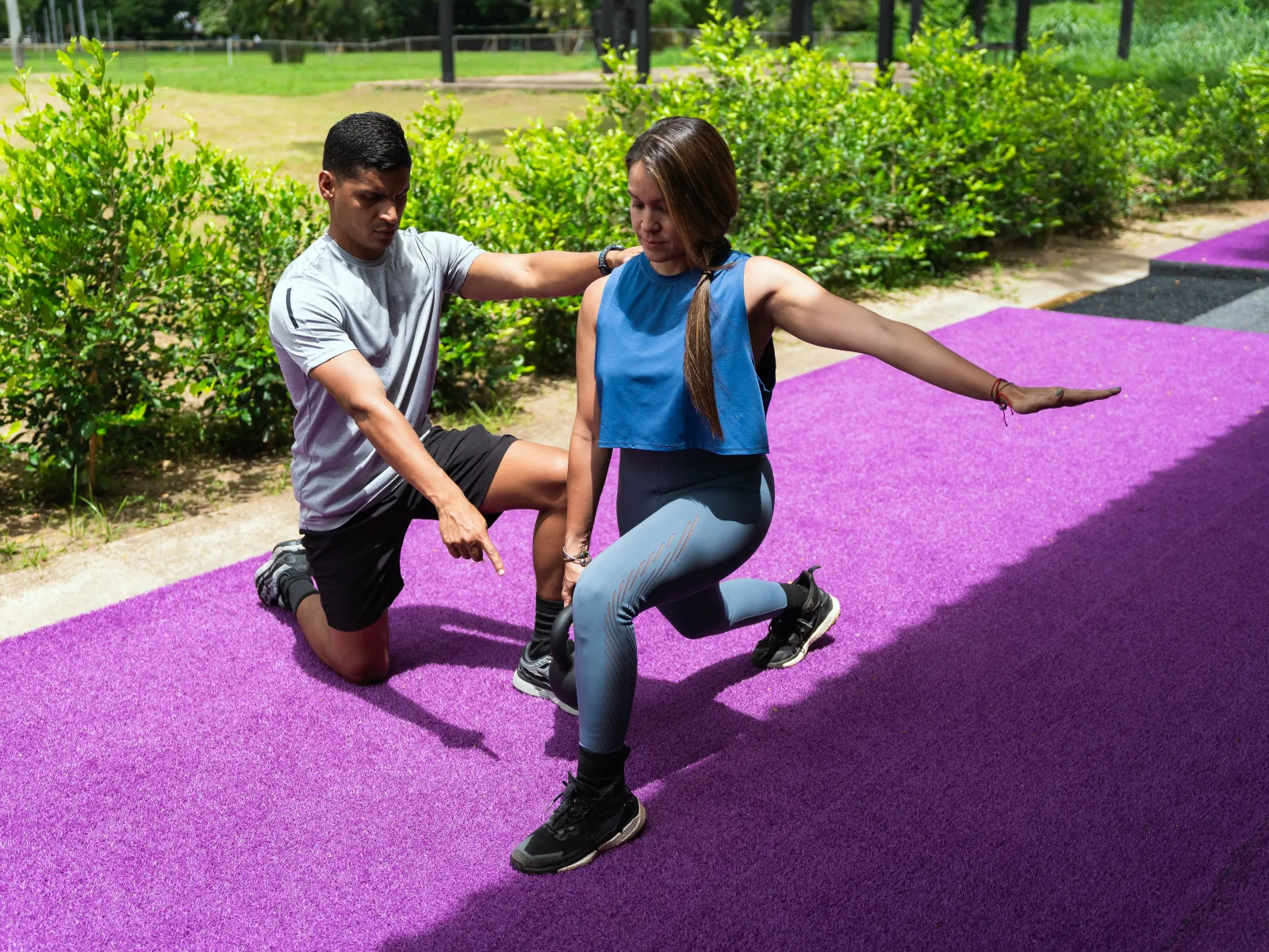 Personal trainer guiding woman through a lunge exercise on purple outdoor gym mat in sunny park.