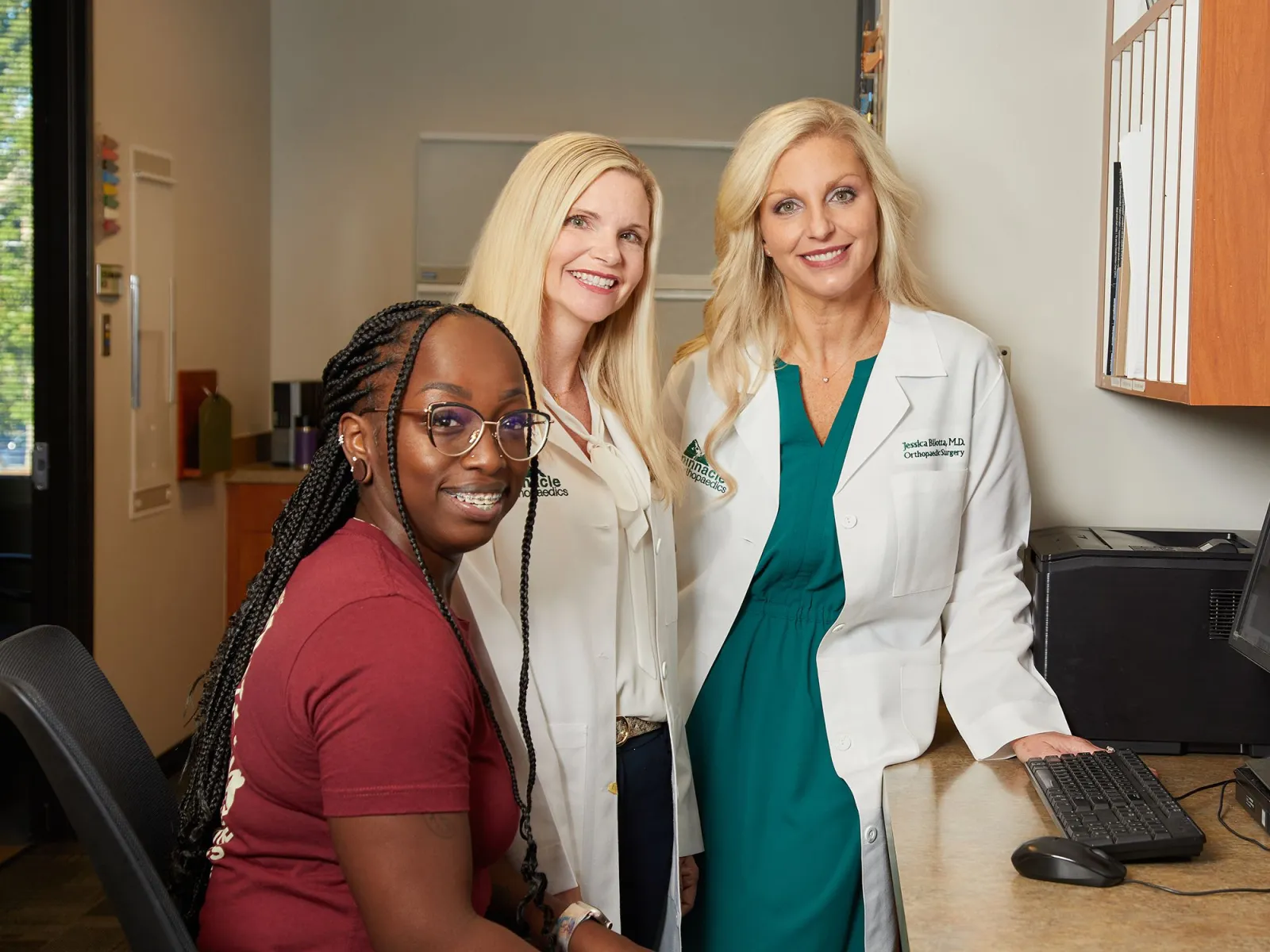 Smiling woman with glasses sitting with two female doctors in white coats at a medical office desk