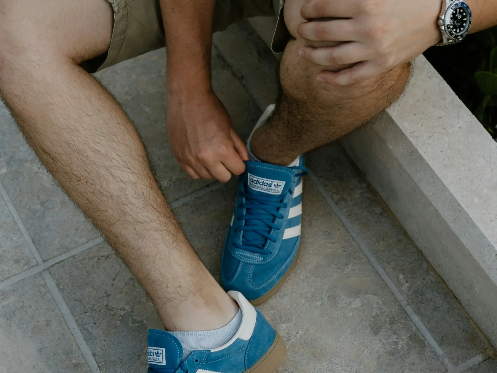 Person wearing beige shorts and blue Adidas sneakers sitting on tiled floor adjusting shoe laces.
