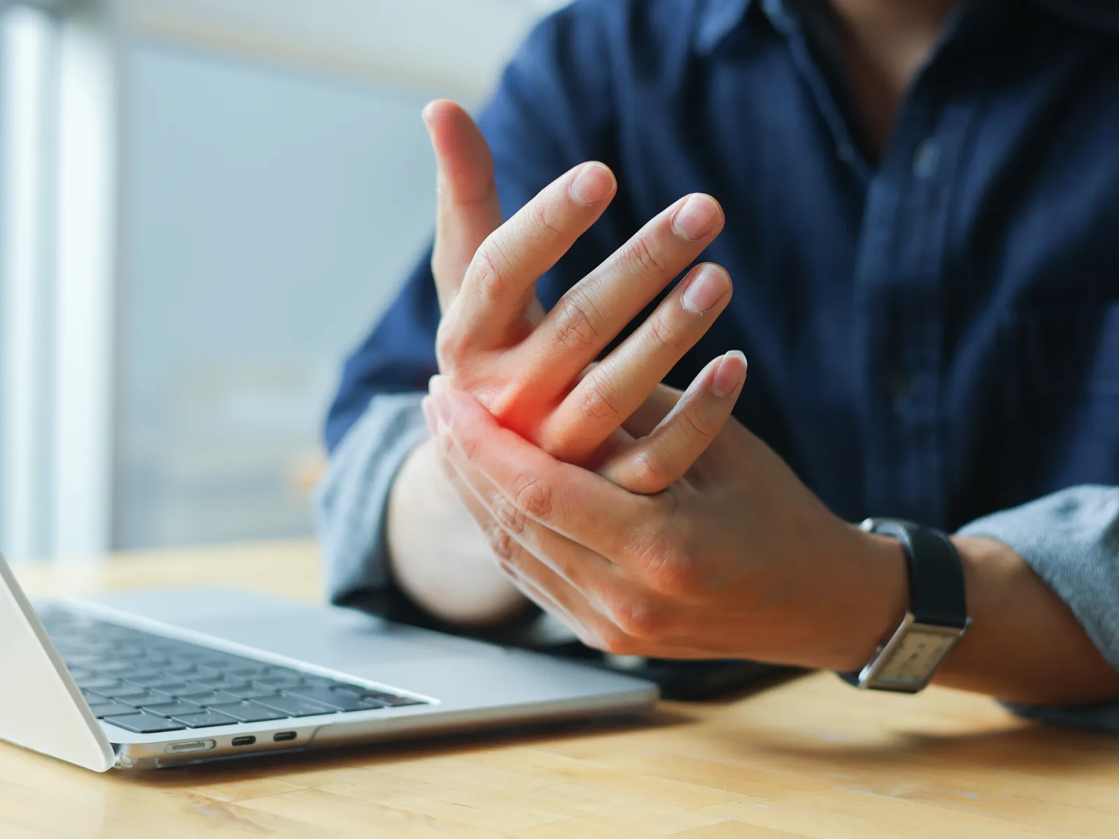 Person sitting at desk with laptop holding wrist in pain or discomfort, highlighting possible carpal tunnel syndrome.