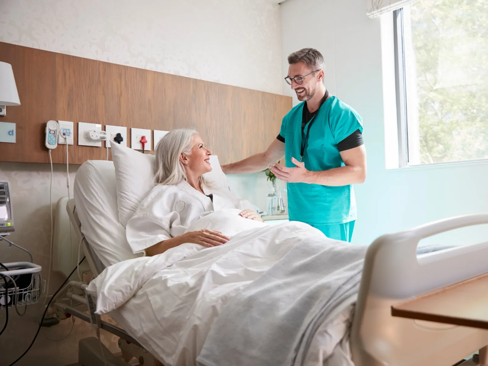 Nurse in teal scrubs talking and smiling with elderly female patient in hospital bed by window