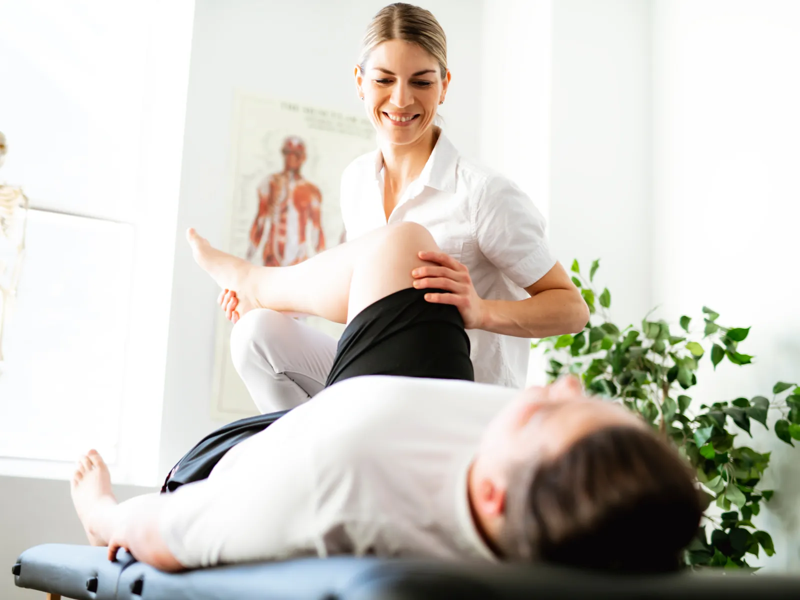 Physiotherapist smiling and stretching patient's leg in a bright clinic with anatomical models and plants