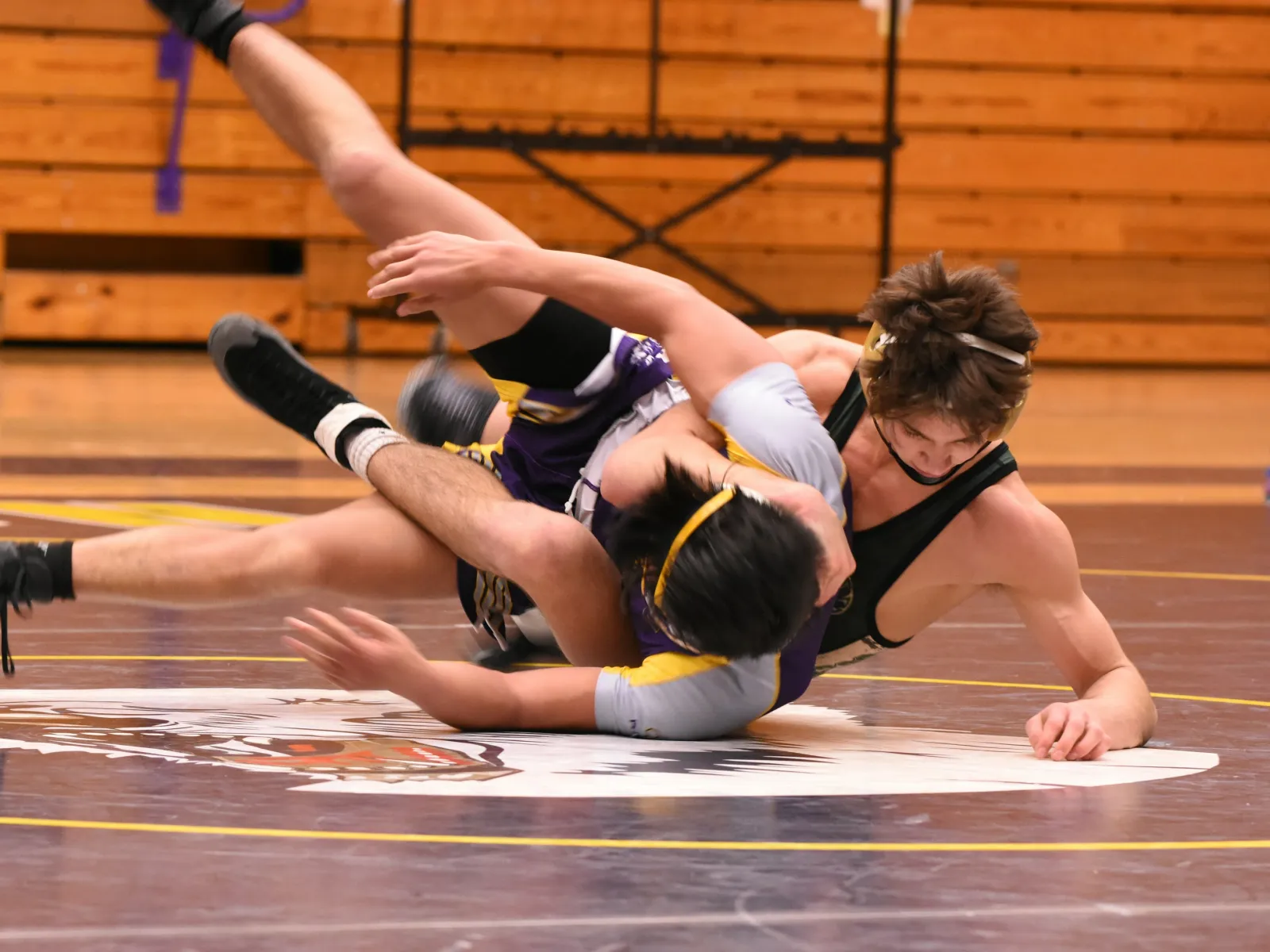 Two high school wrestlers grapple intensely on a gymnasium mat during a match inside a school gym.