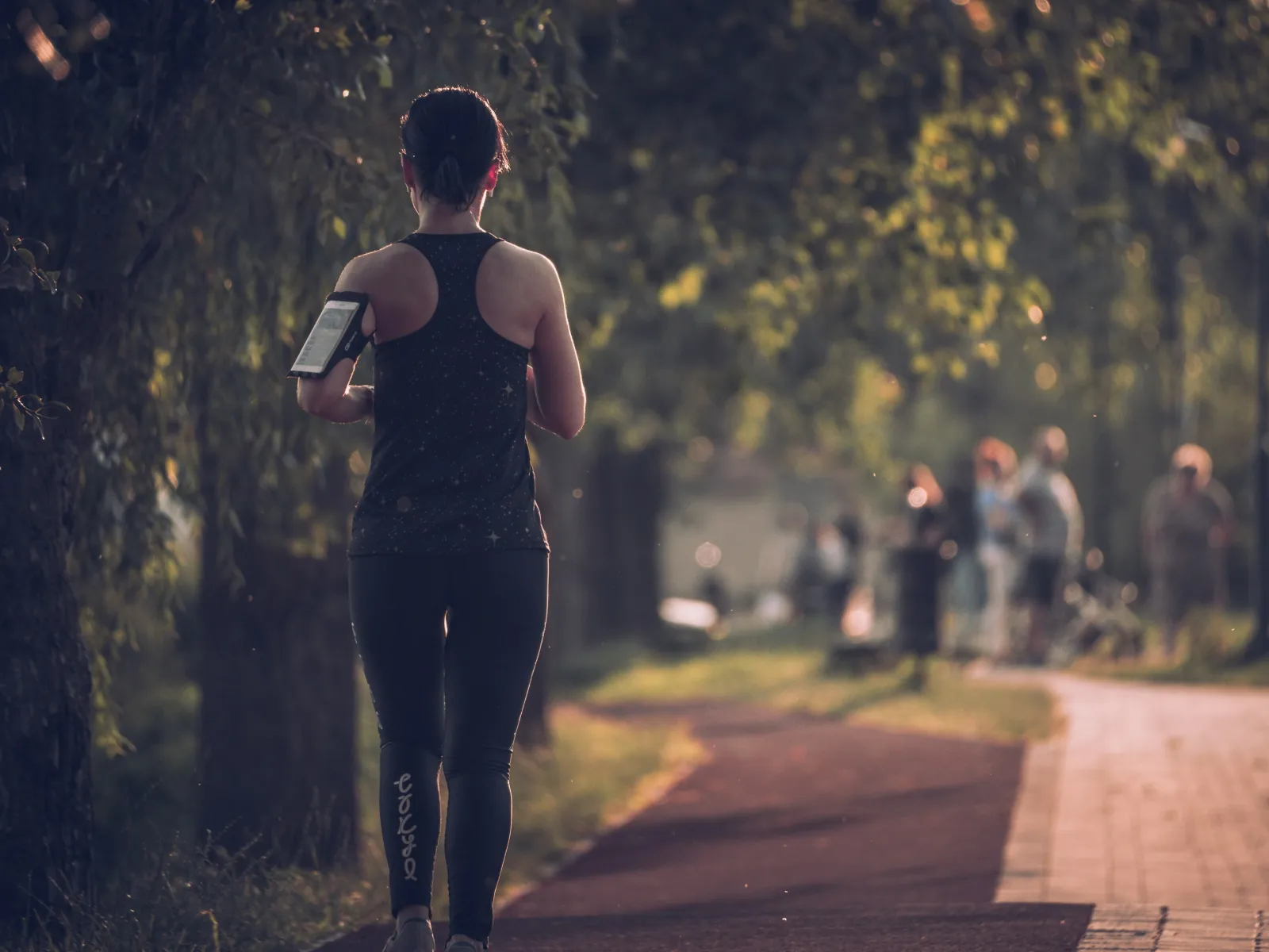 Woman jogging on a tree-lined path wearing black athletic wear with blurred park visitors in the background.