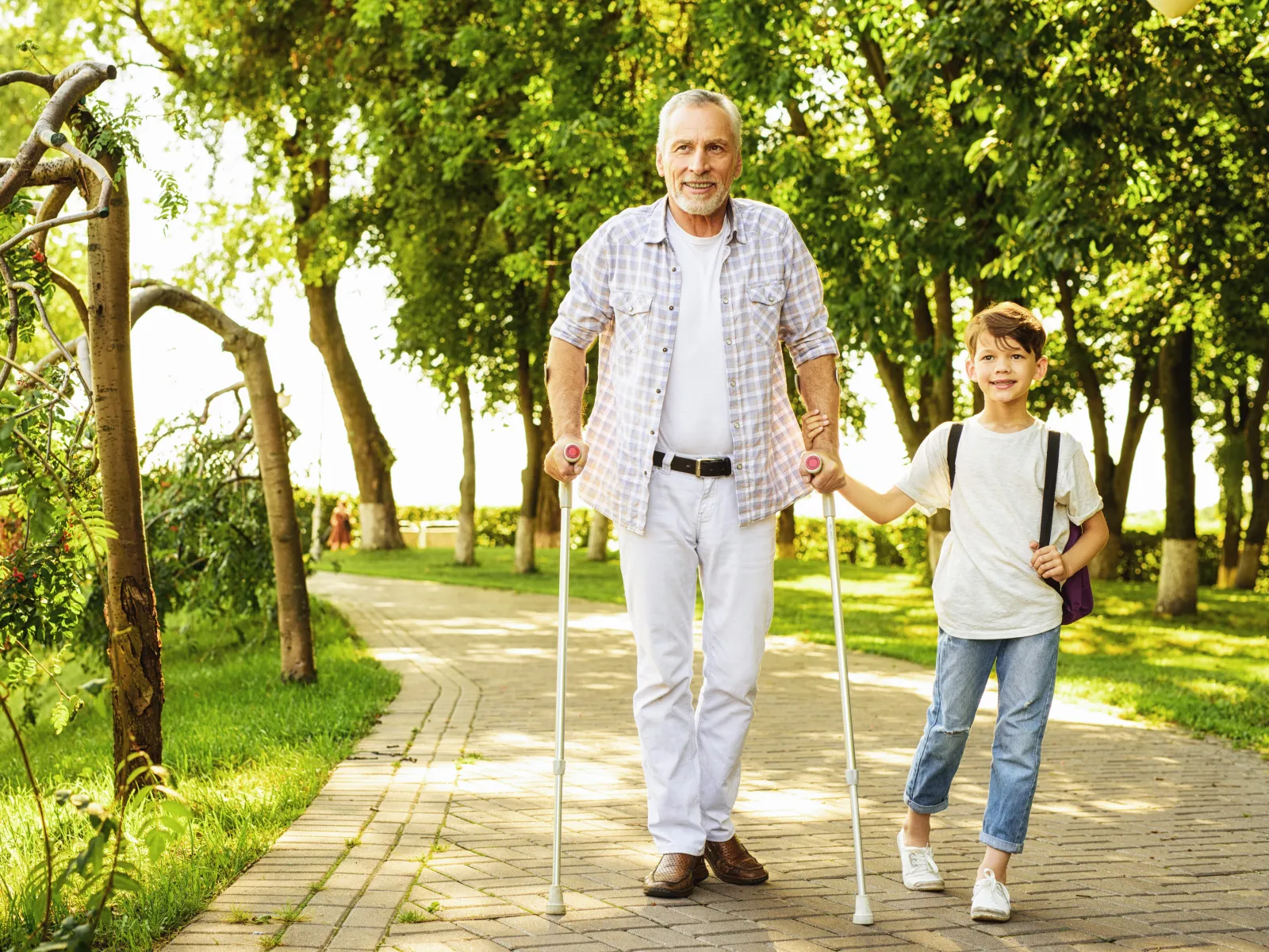 Senior man with crutches walking hand in hand with young boy on a sunny park pathway