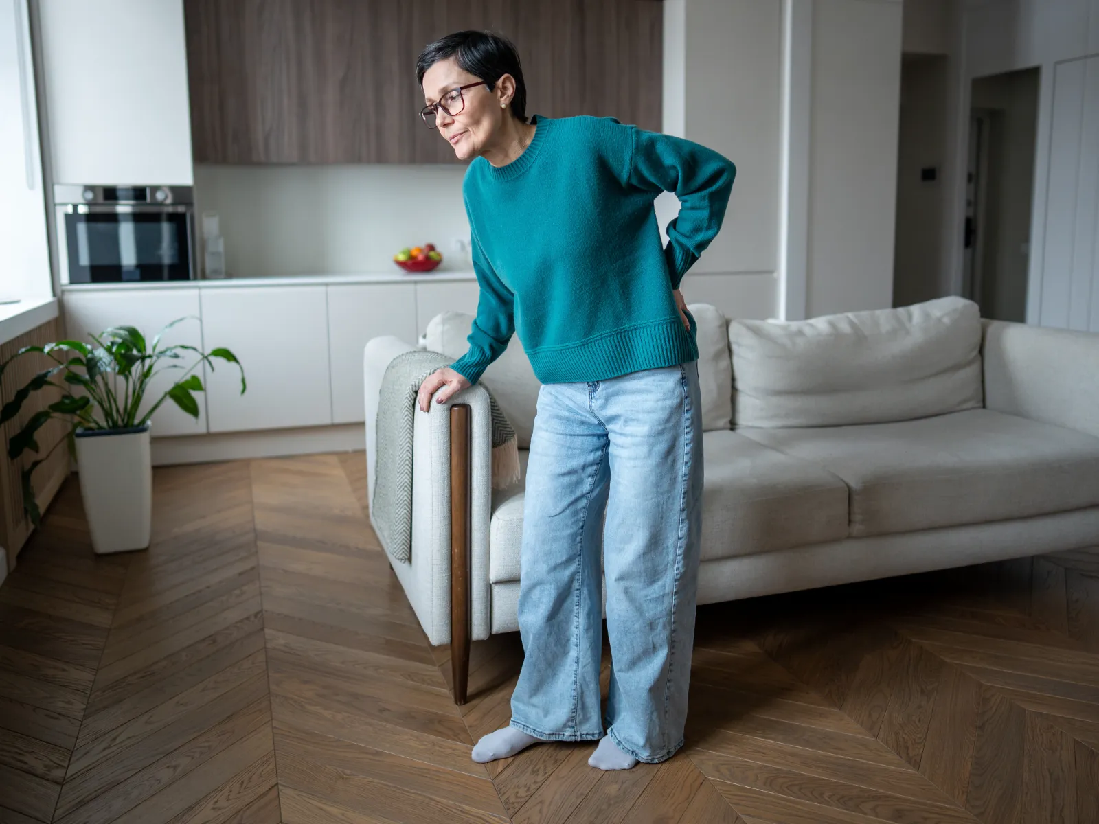 Woman in teal sweater and jeans holding her lower back, standing in a modern living room with a beige sofa.