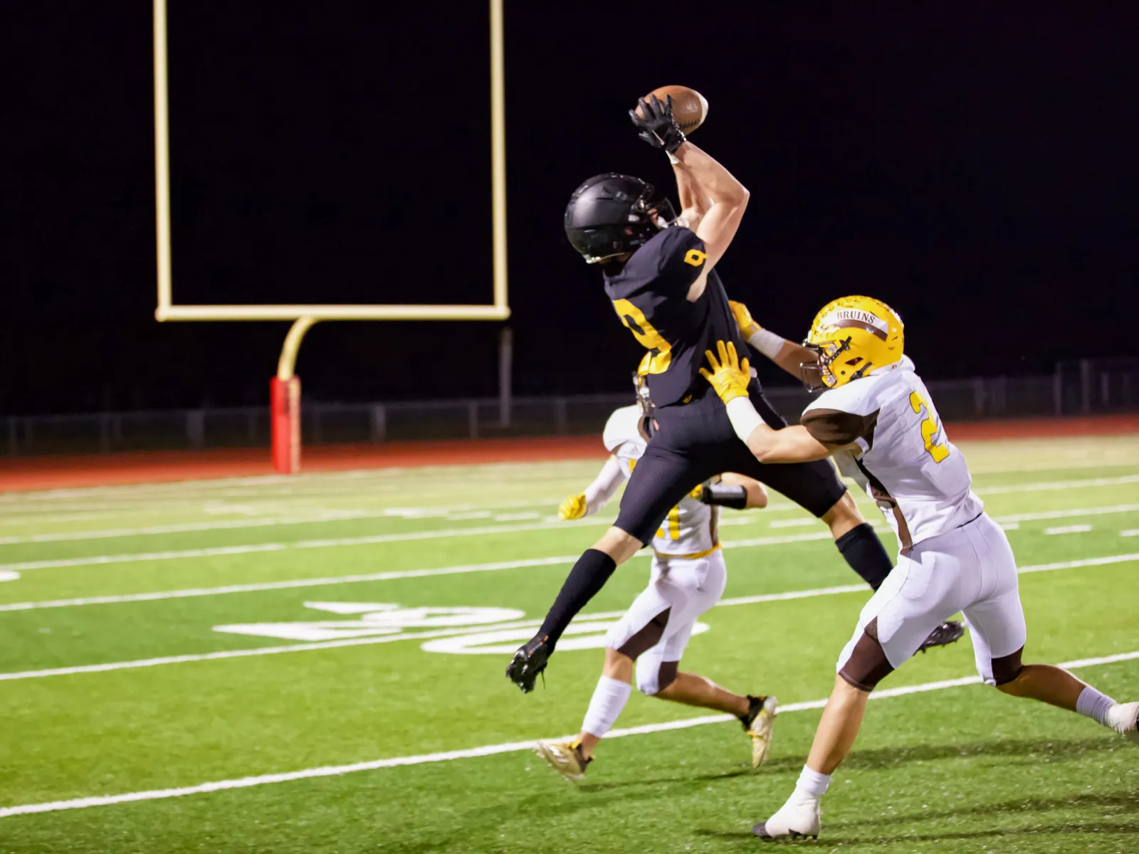 Football player in black jersey jumps to catch ball while defended by a player in white and yellow uniform on field.