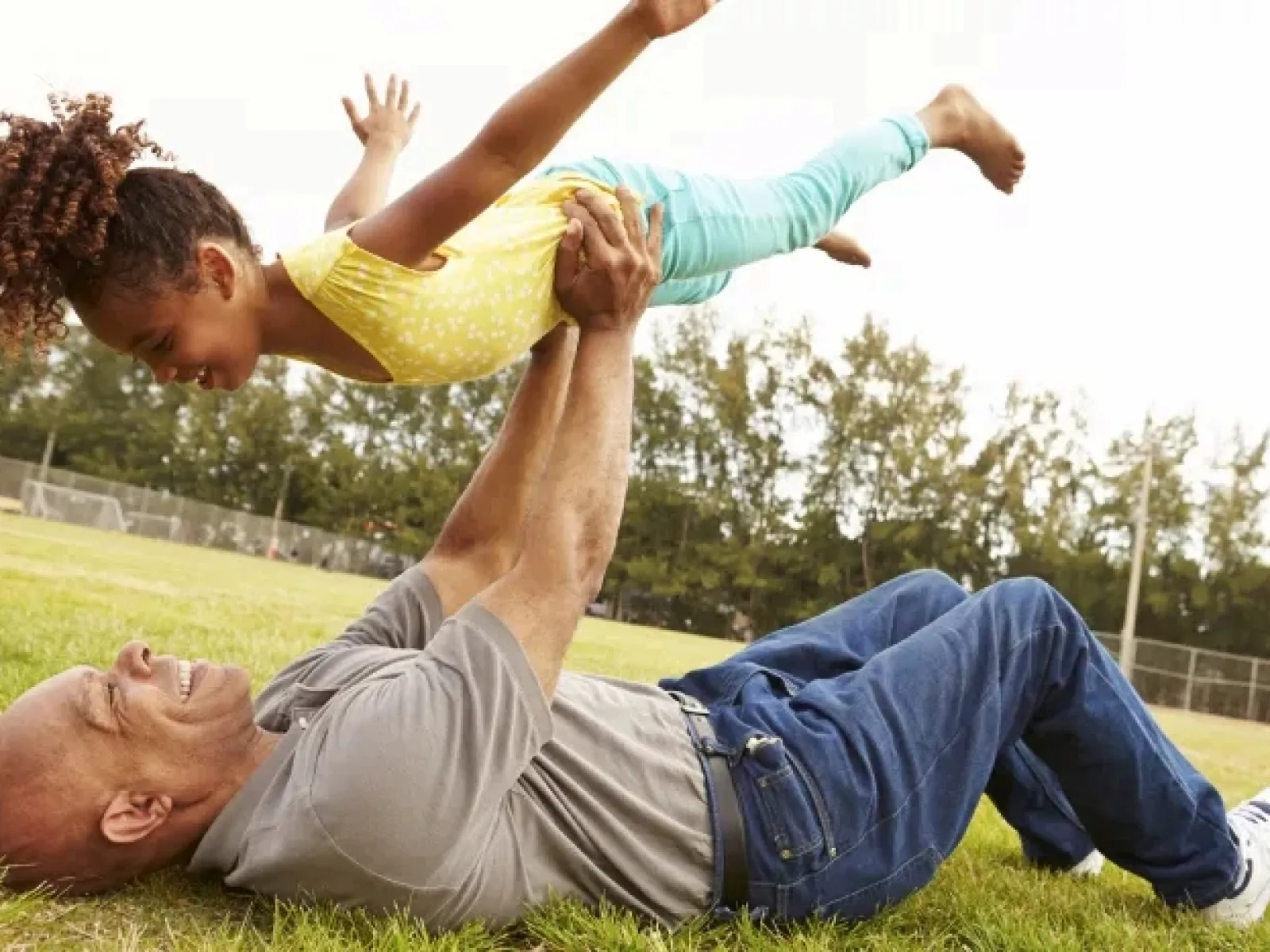 Grandfather lying on grass lifting smiling granddaughter in the air in a sunny park outdoors