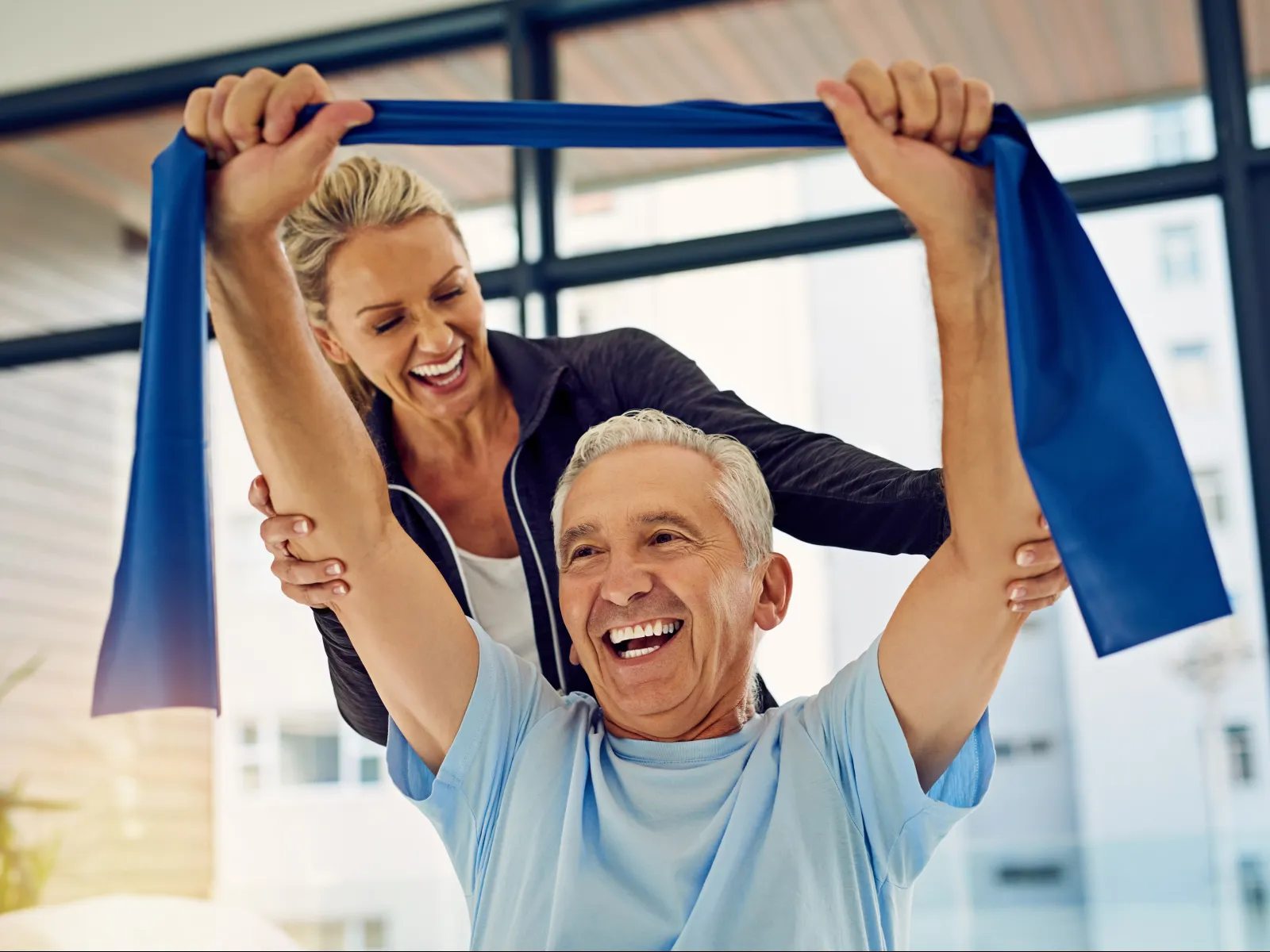 Senior man exercising with resistance band assisted by smiling female trainer in bright gym.