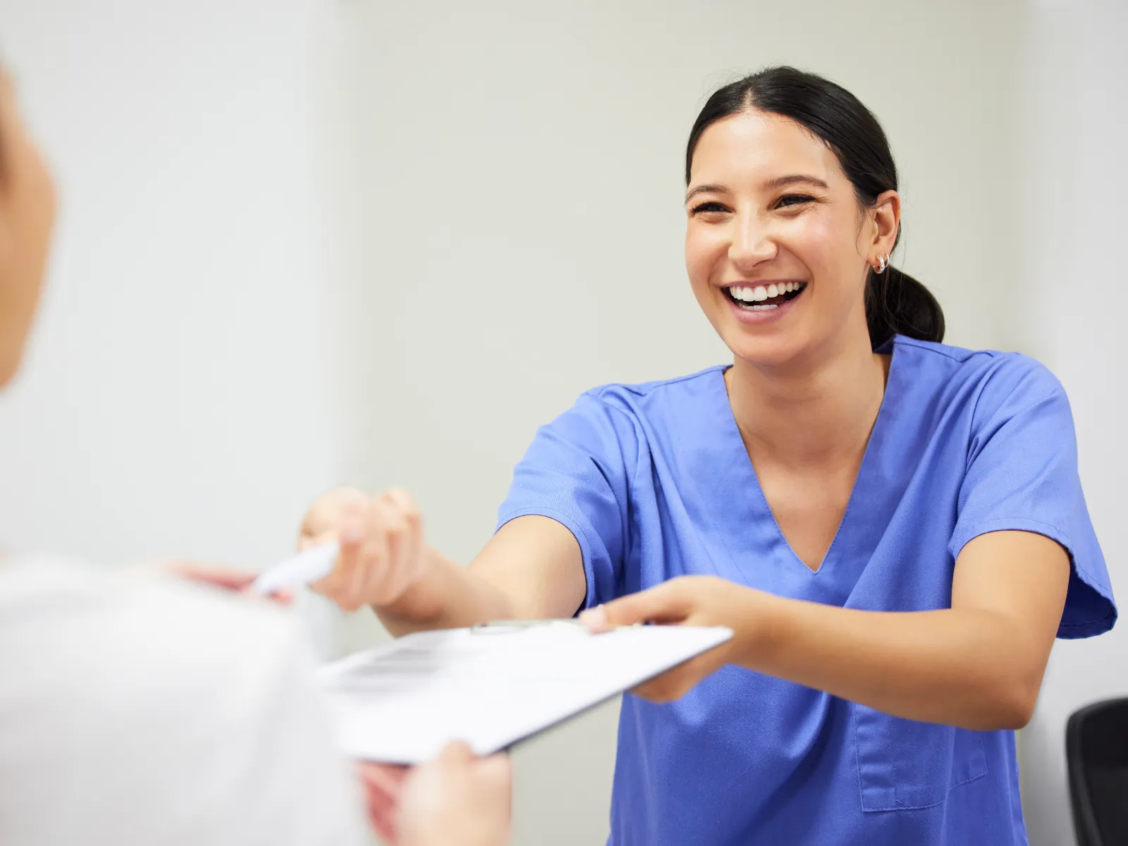 Smiling healthcare worker in blue scrubs handing clipboard to patient in bright medical office.