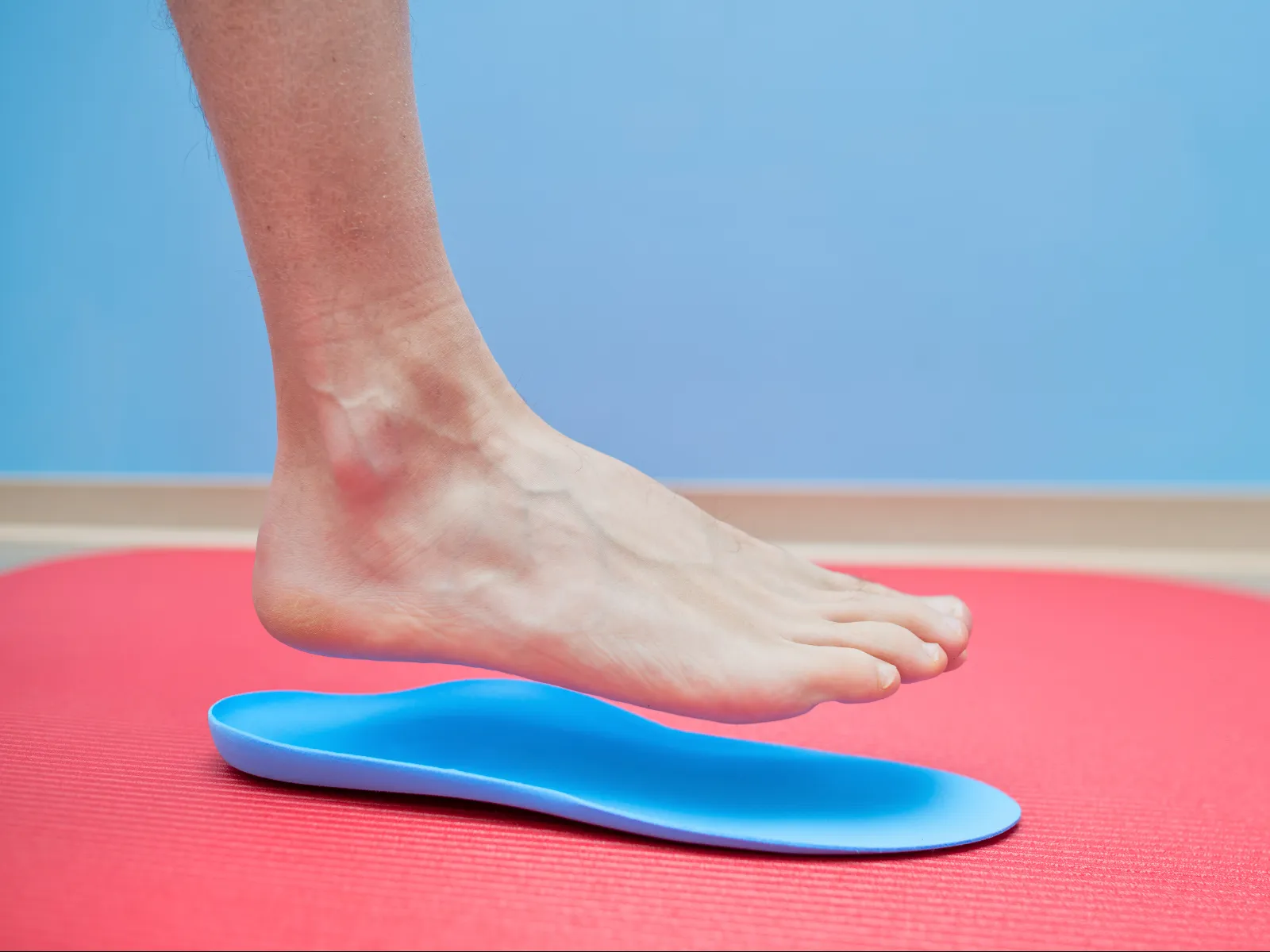Bare foot hovering above a blue orthotic insole on a red yoga mat against a blue background