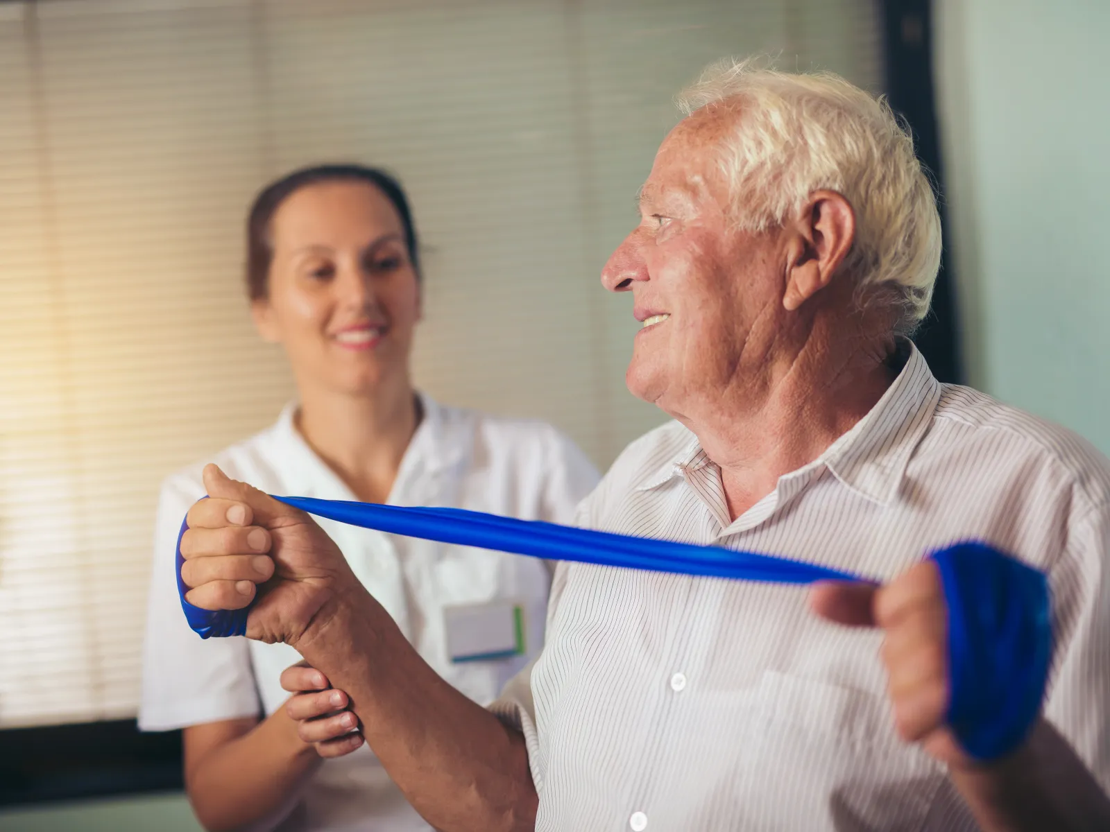 Elderly man exercises with blue resistance band guided by female therapist in rehabilitation setting
