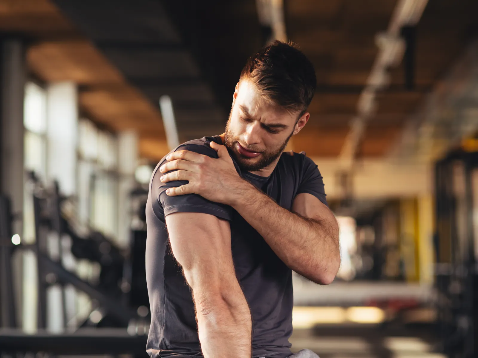 Man in gym wearing black shirt holding his shoulder in pain, muscle strain or injury concept