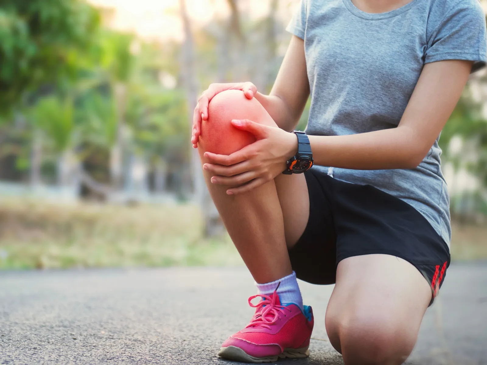 Person kneeling on pavement holding painful knee outdoors, wearing pink shoes and gray shirt during exercise.