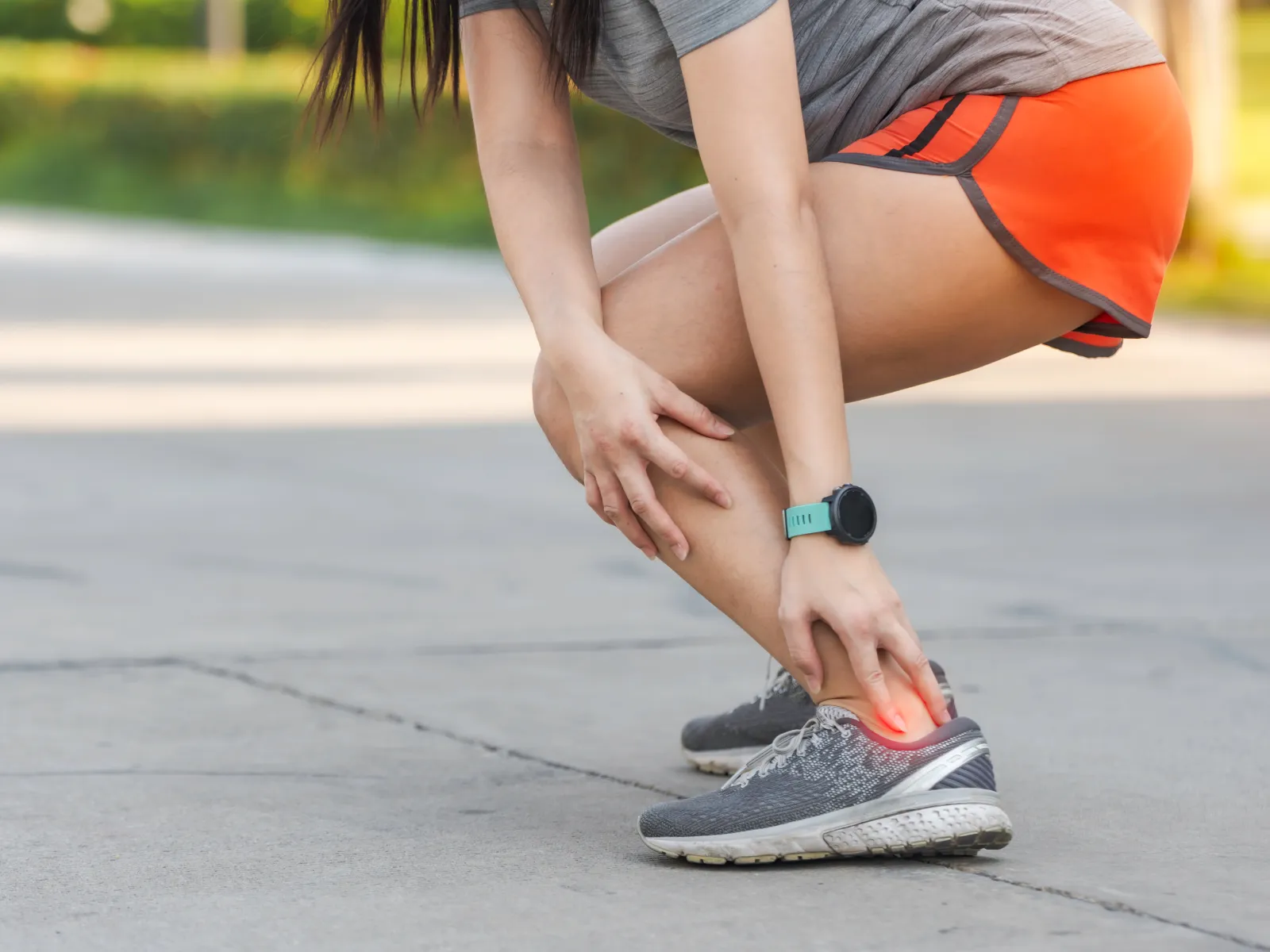 Runner in orange shorts holding aching ankle outdoors on pavement, highlighting ankle pain or injury.
