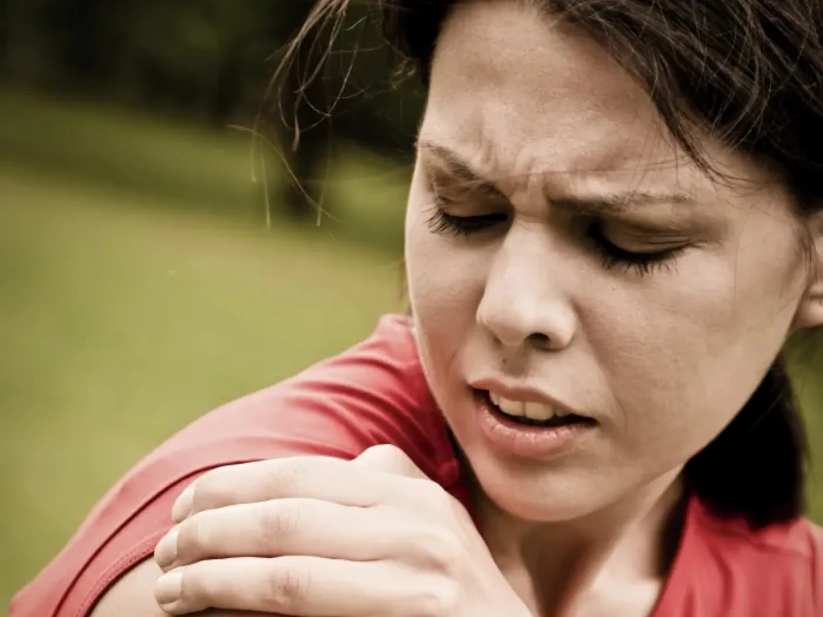 Woman in red shirt holding shoulder in pain, outdoors with blurred green background.