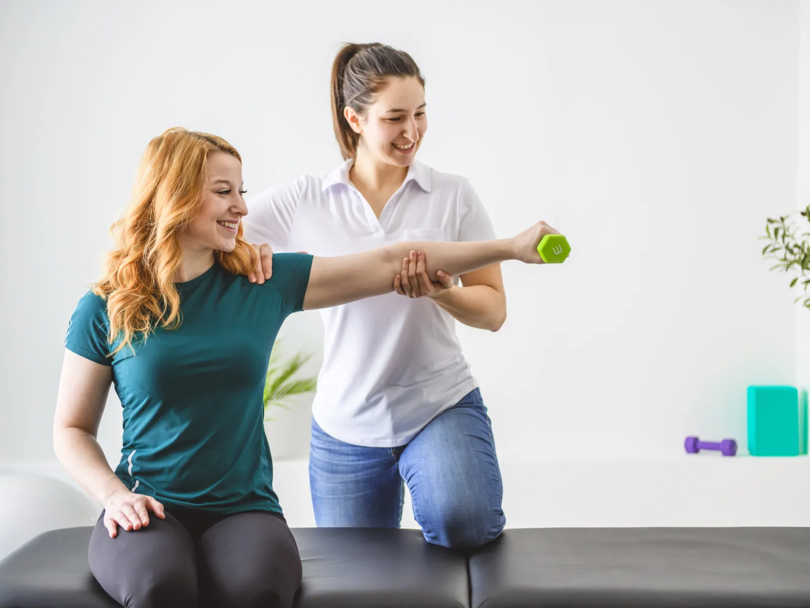 Physical therapist assisting woman with arm exercise using a small green dumbbell in a bright therapy room.