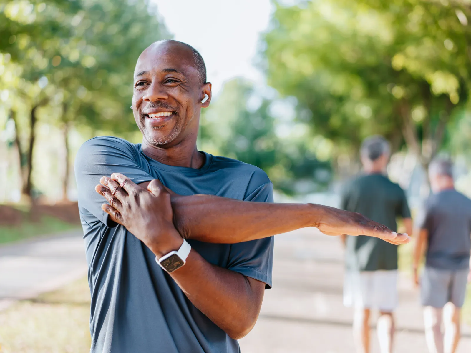 Smiling man stretches arm outdoors on a sunny day wearing wireless earbuds and a fitness watch.