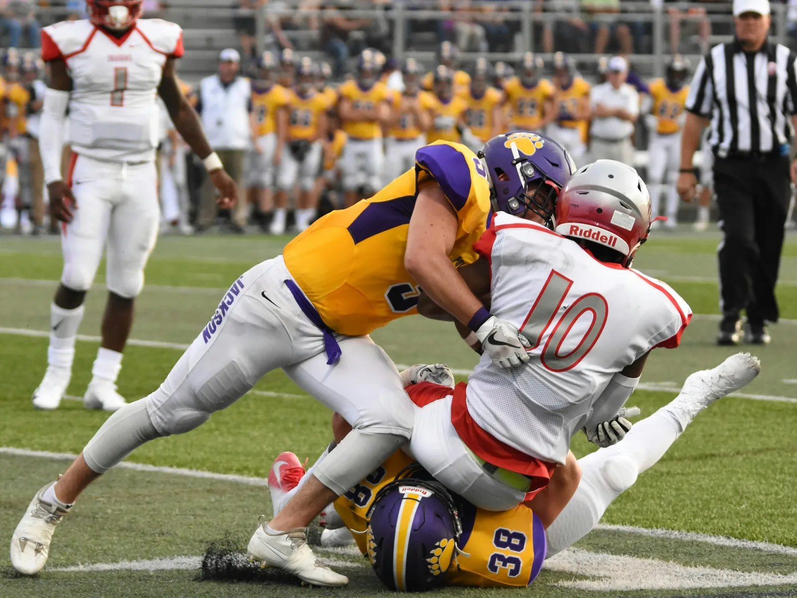 Football player in white jersey tackled by two players in yellow and purple uniforms during a game.