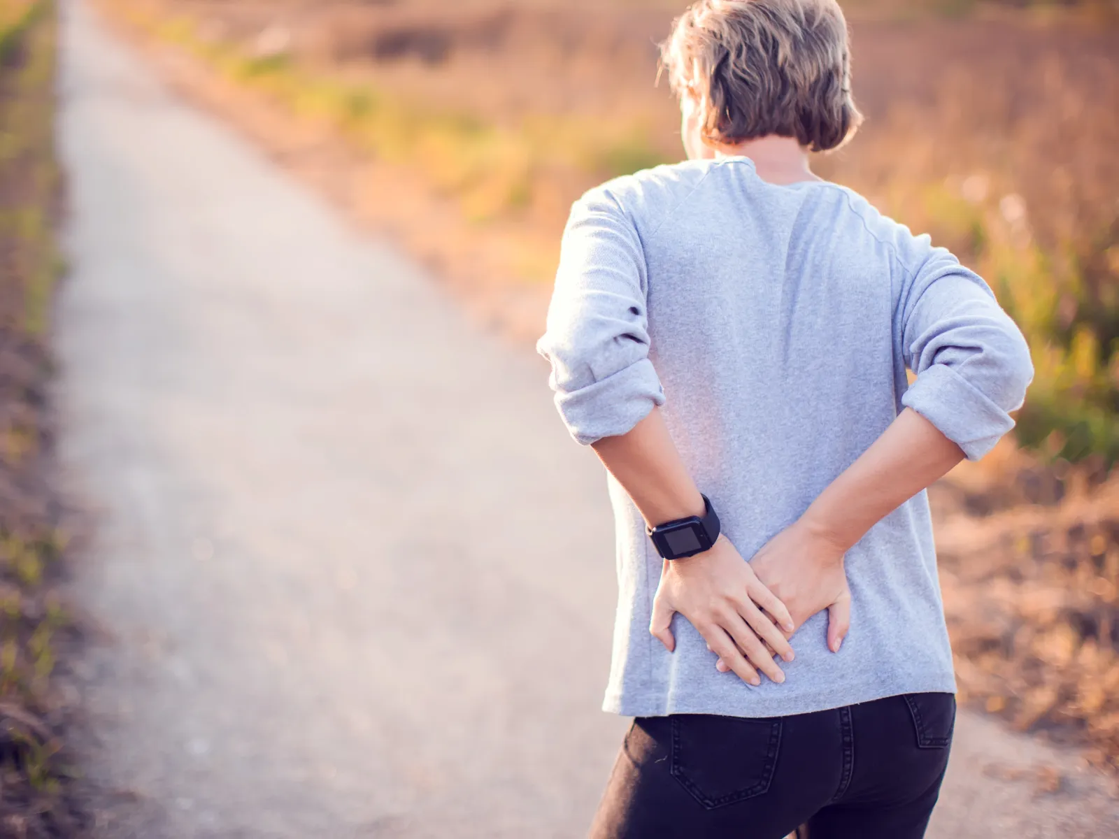 Middle-aged person holding lower back in pain while standing on an outdoor path with autumn surroundings