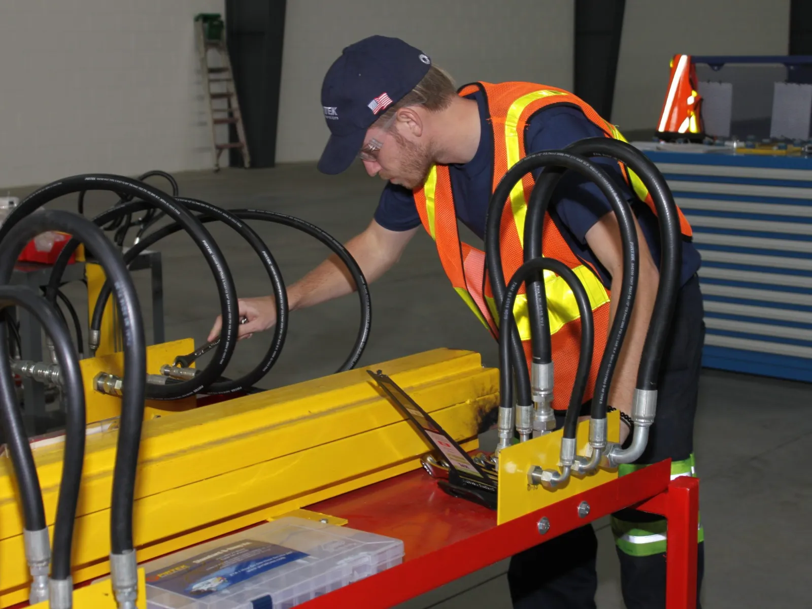 Worker in safety vest inspects industrial equipment with hydraulic hoses in a warehouse setting.
