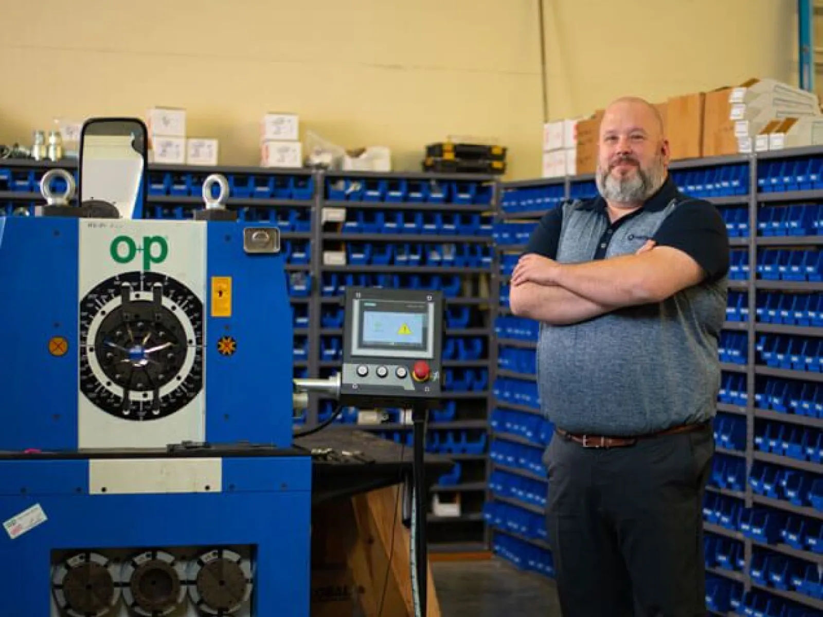 Man standing next to large blue industrial machinery with storage bins in the background in a workshop.