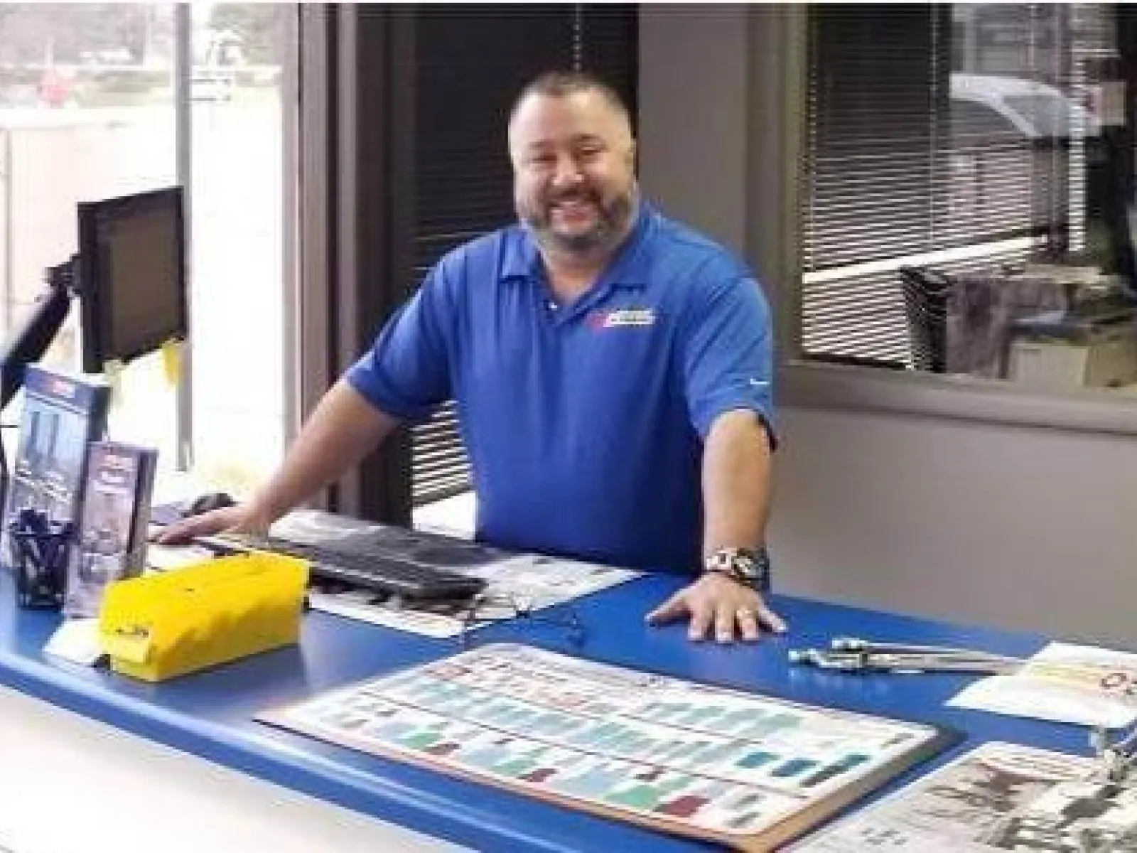 Smiling man in blue shirt stands behind office counter with brochures near large window showing parked service van outside.