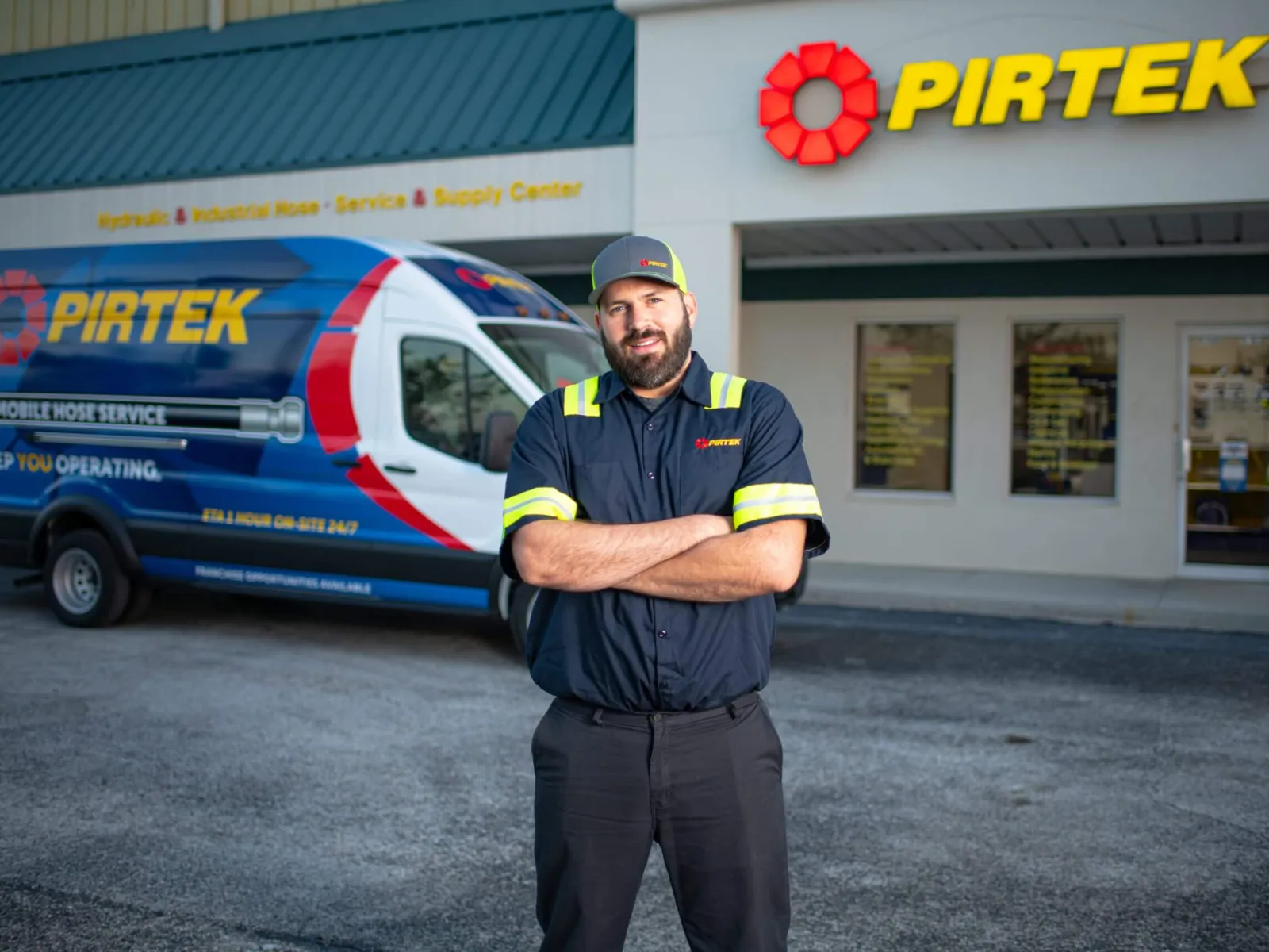 Pirtek technician in uniform stands with arms crossed outside service center next to branded service van.