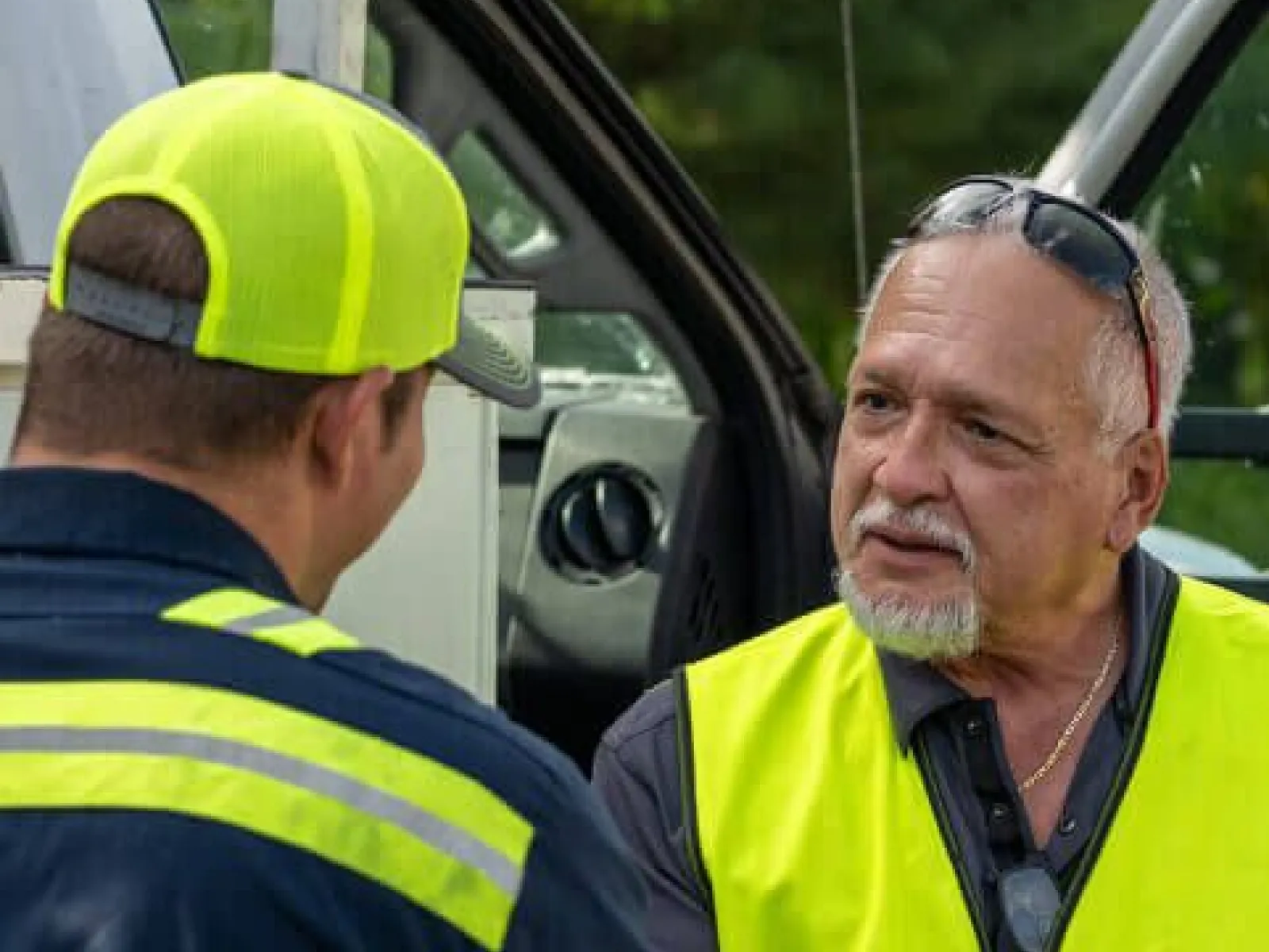 Two construction workers wearing safety vests shaking hands near a work vehicle outdoors