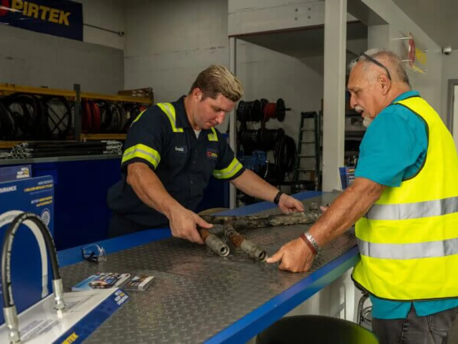 Two men examining metal pipes on a workshop counter with PIRTEK branding in the background.