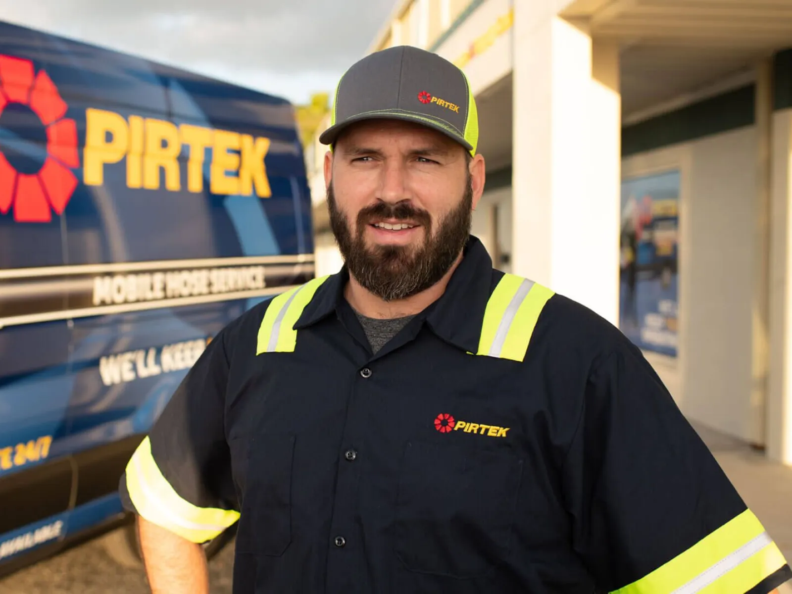 Man in high-visibility workwear and cap stands in front of a Pirtek mobile hose service van outdoors.