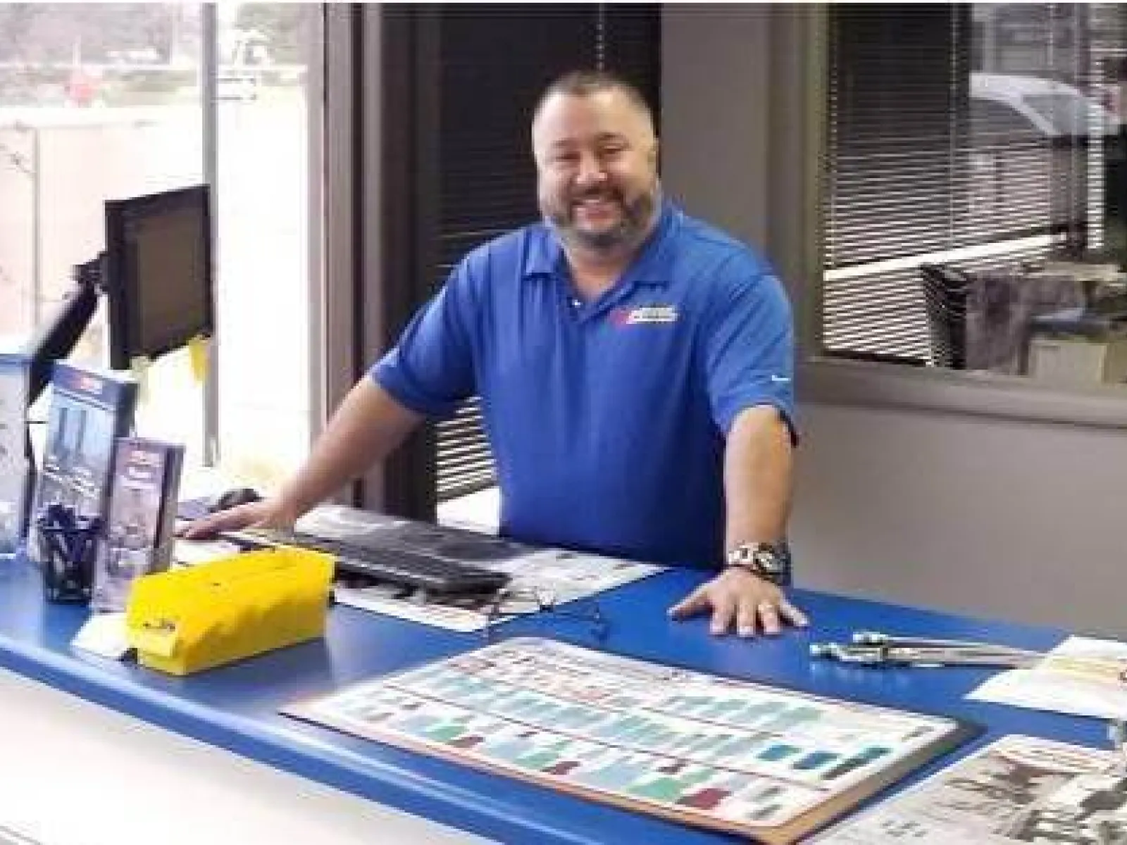 Smiling employee in blue uniform stands behind showroom counter with tools and brochures, van visible outside window
