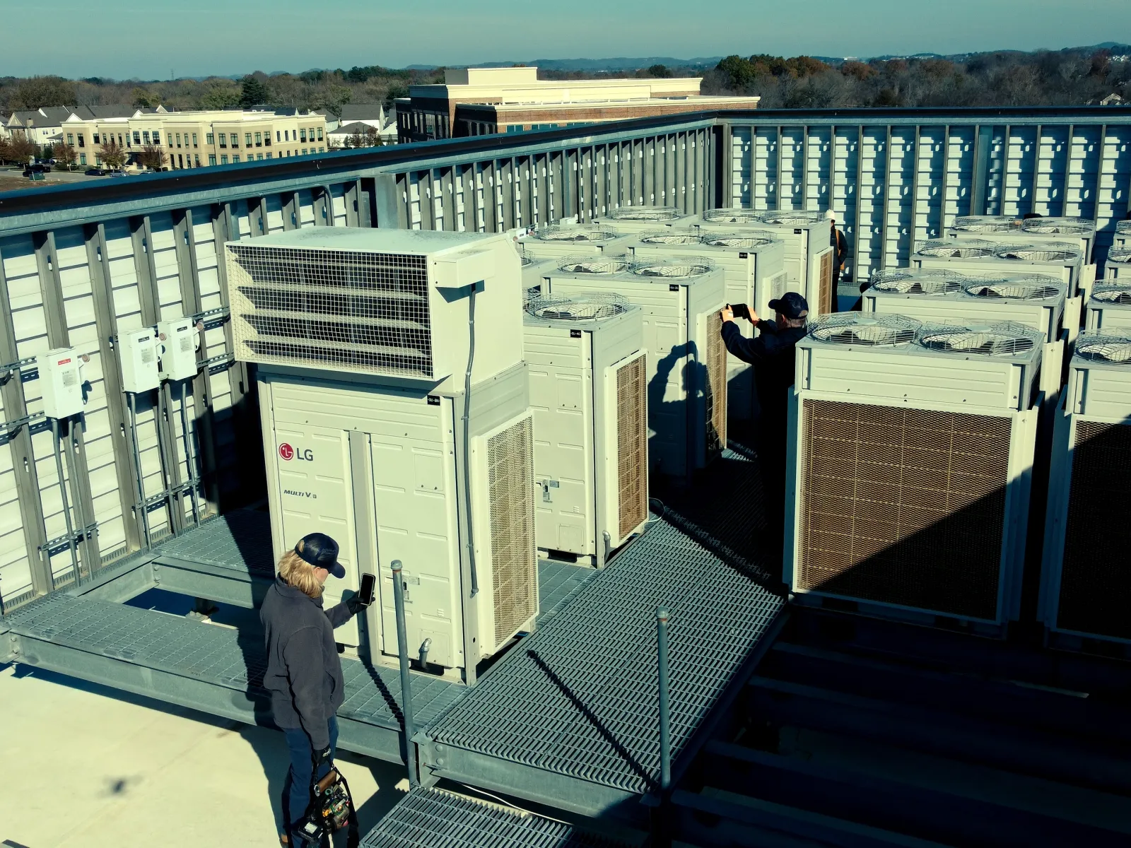 Technicians inspecting large LG HVAC units on a rooftop with clear skies and distant buildings.