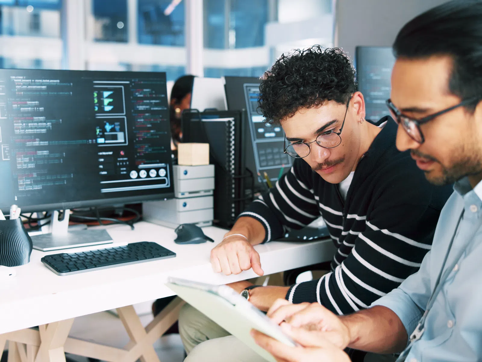 Two male software developers collaborating at a workstation with multiple monitors displaying code.