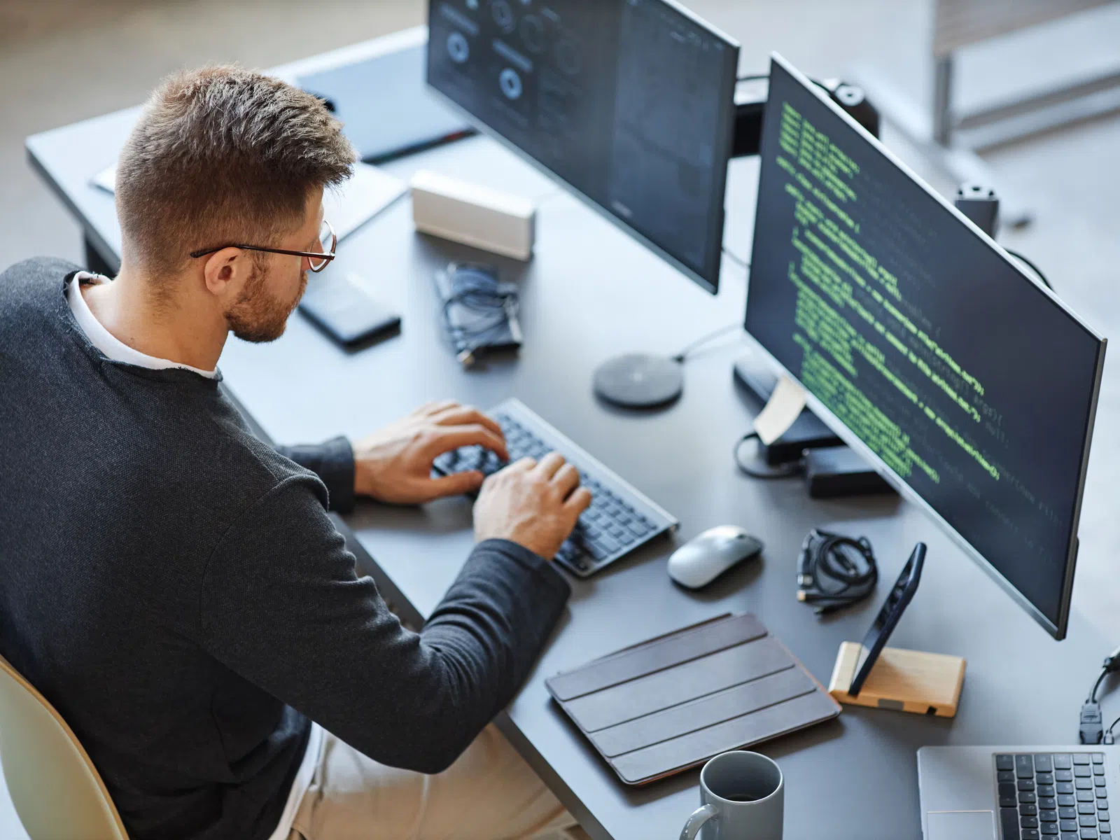 Man typing code on keyboard with dual monitors displaying programming and workspace items on desk.