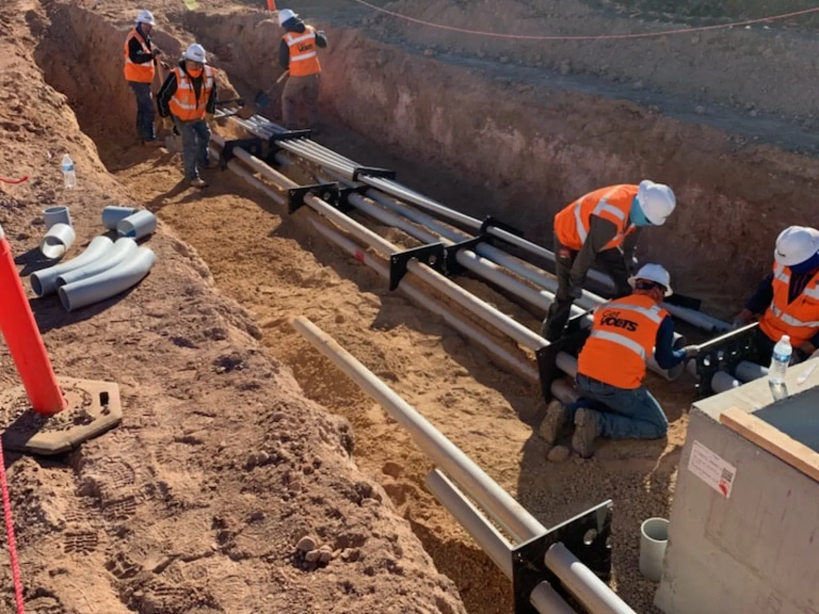 Construction workers in orange vests installing underground pipes in a trench near a metal fence under sunlight.