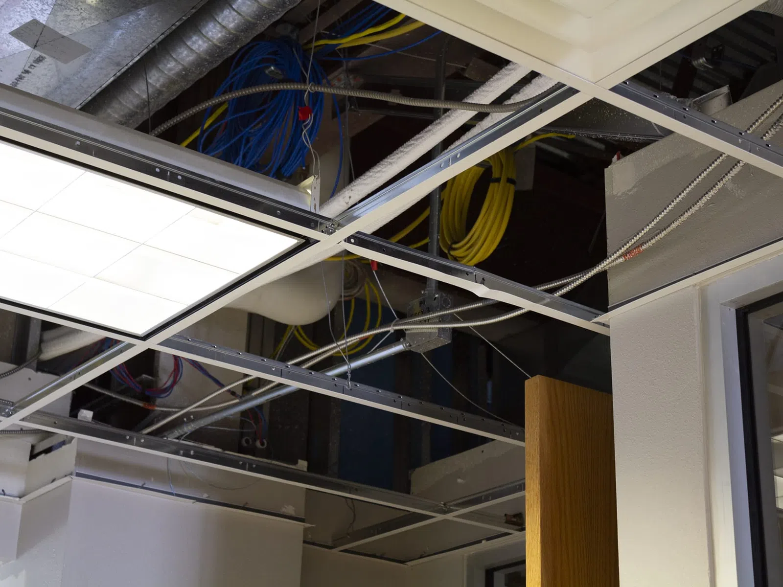 Exposed ceiling framework with electrical wiring, ventilation ducts, and a bright ceiling light panel in an office space.