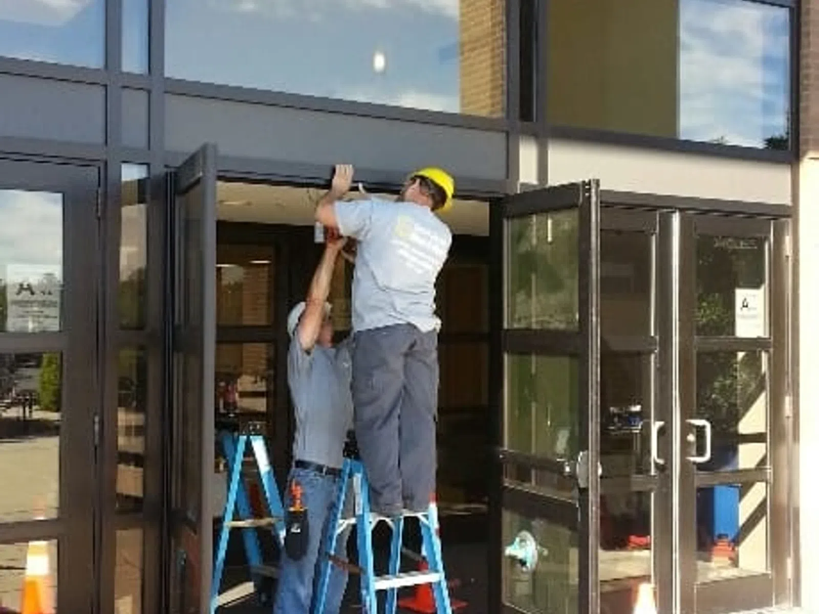 Two workers install or repair a glass entrance door at a commercial building using ladders and safety cones.