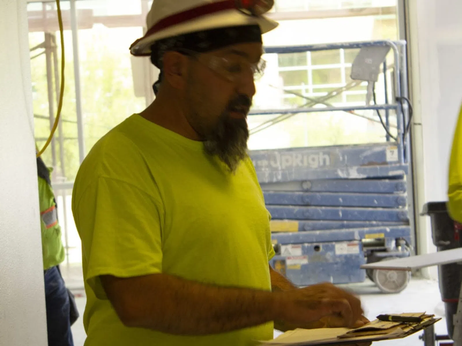 Construction worker in a bright yellow shirt and hard hat reviewing documents on a clipboard indoors.